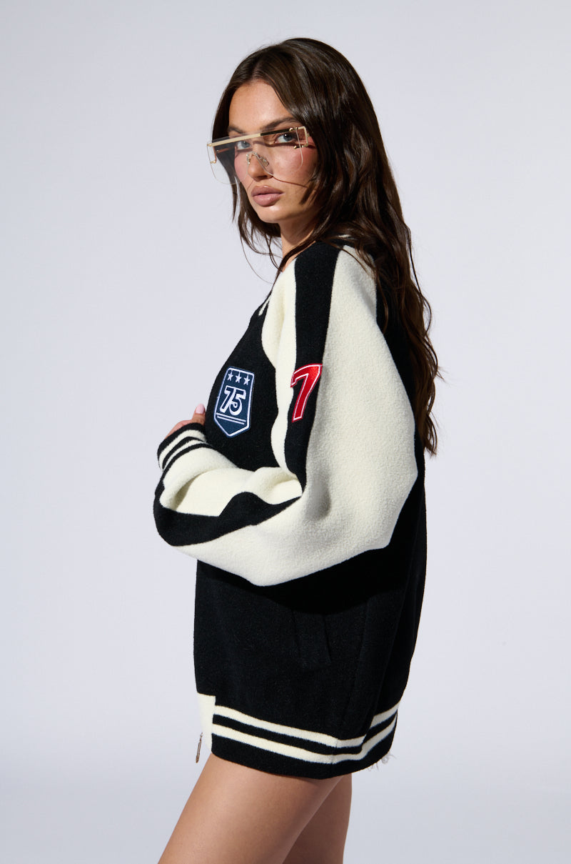 A woman with long brown hair wears an oversized DOWN TIME KNIT BOMBER from the AKIRA Label, featuring a black and white color block pattern and adorned with patches, including one that says "75" on the front and another red "7" on the sleeve. She has transparent glasses with a gold frame and stands against a plain white background.