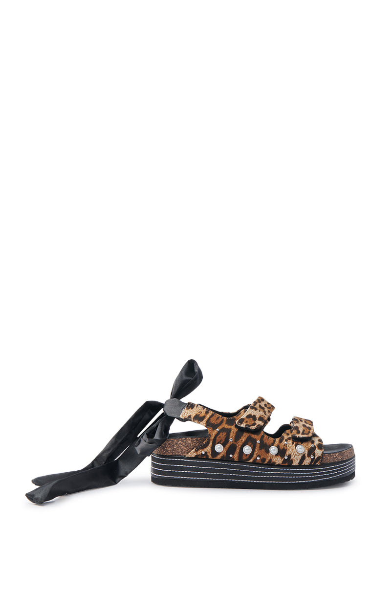 The AKIRA MACKLEY LEOPARD FLATFORM SANDAL, shown against a white background, features an open almond toe, two thick straps, and a heel satin lace tie. Its chic design is enhanced by a sole layered with black and white stripes.
