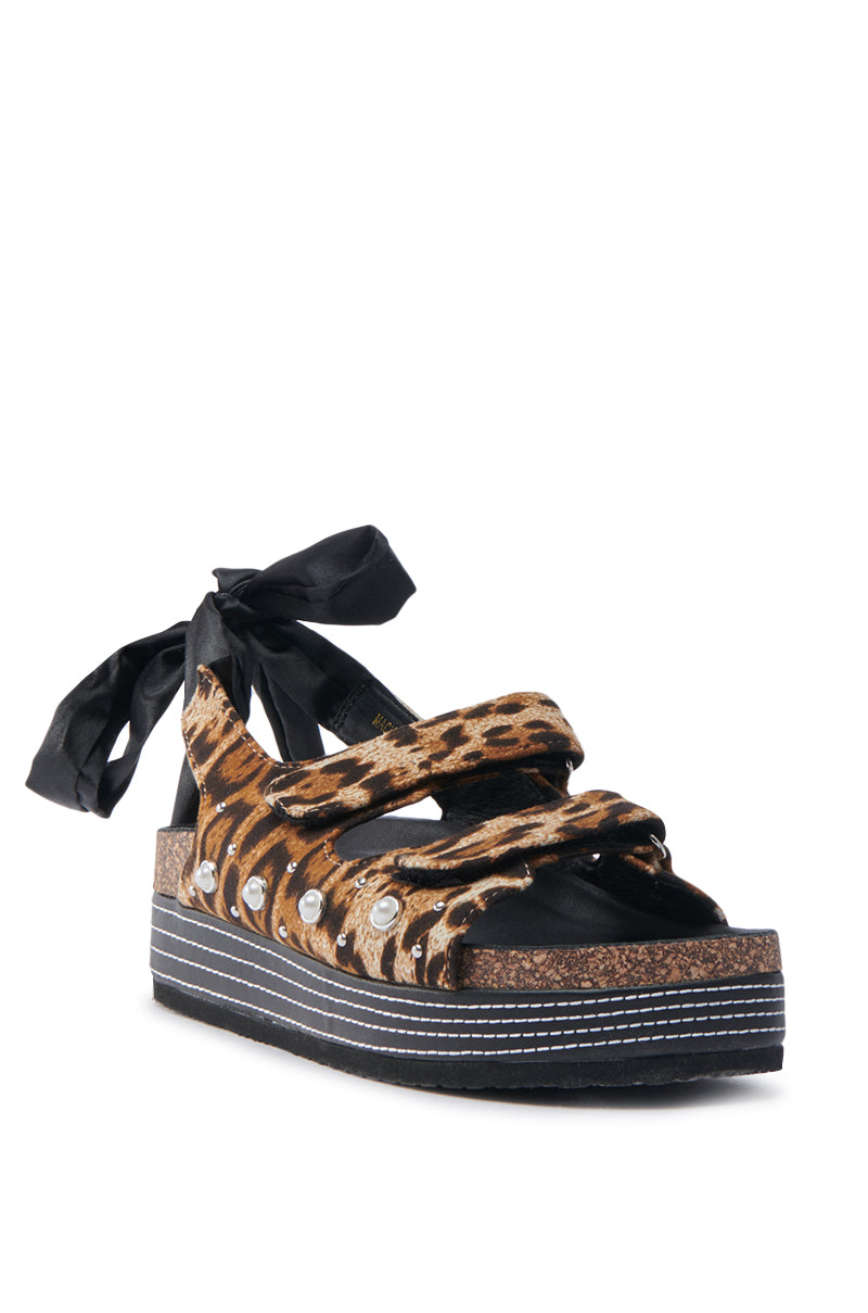 The AKIRA MACKLEY LEOPARD FLATFORM SANDAL showcases animal print straps with metal studs, chic black ribbon ties, and a thick black sole with white stitching. Crafted from faux leather for style and comfort.