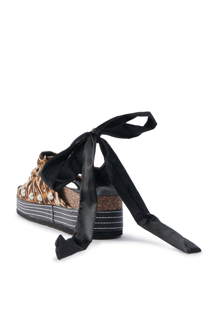 The AKIRA MACKLEY LEOPARD FLATFORM SANDAL features a bold leopard print, large black ribbon ties at the back, faux leather finish, chunky sole with stitching details, and a textured footbed for an unconventional style statement.