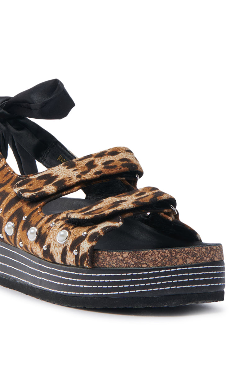 Close-up of AKIRA MACKLEY LEOPARD FLATFORM SANDAL, featuring a leopard design with an open almond toe, faux leather flatform, two straps with silver grommets, black ribbon tie at the back, and a cork-patterned sole accented with black and white stripes.