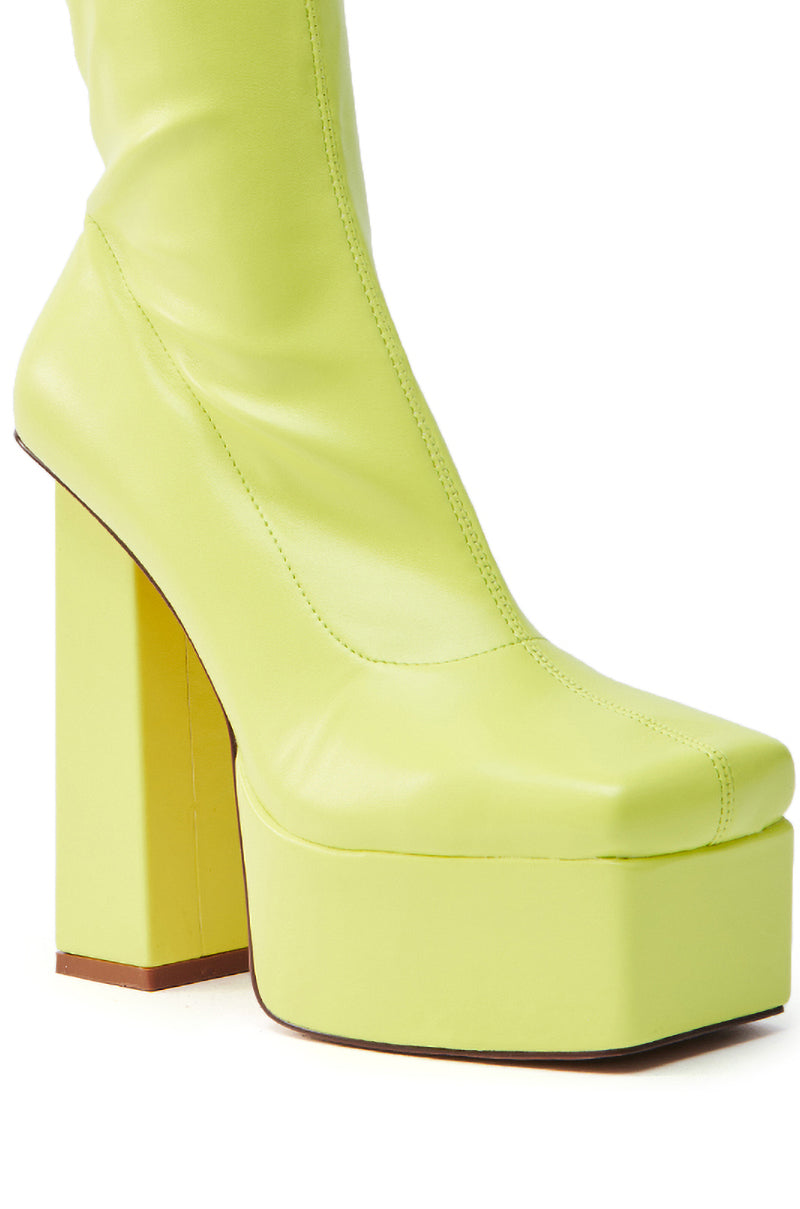 A close-up of the AZALEA WANG MADDER LIME PLATFORM BOOTIE shows a faux leather shoe featuring a chunky high heel, square toe, and smooth mid-calf design. The brown sole enhances its striking lime color.