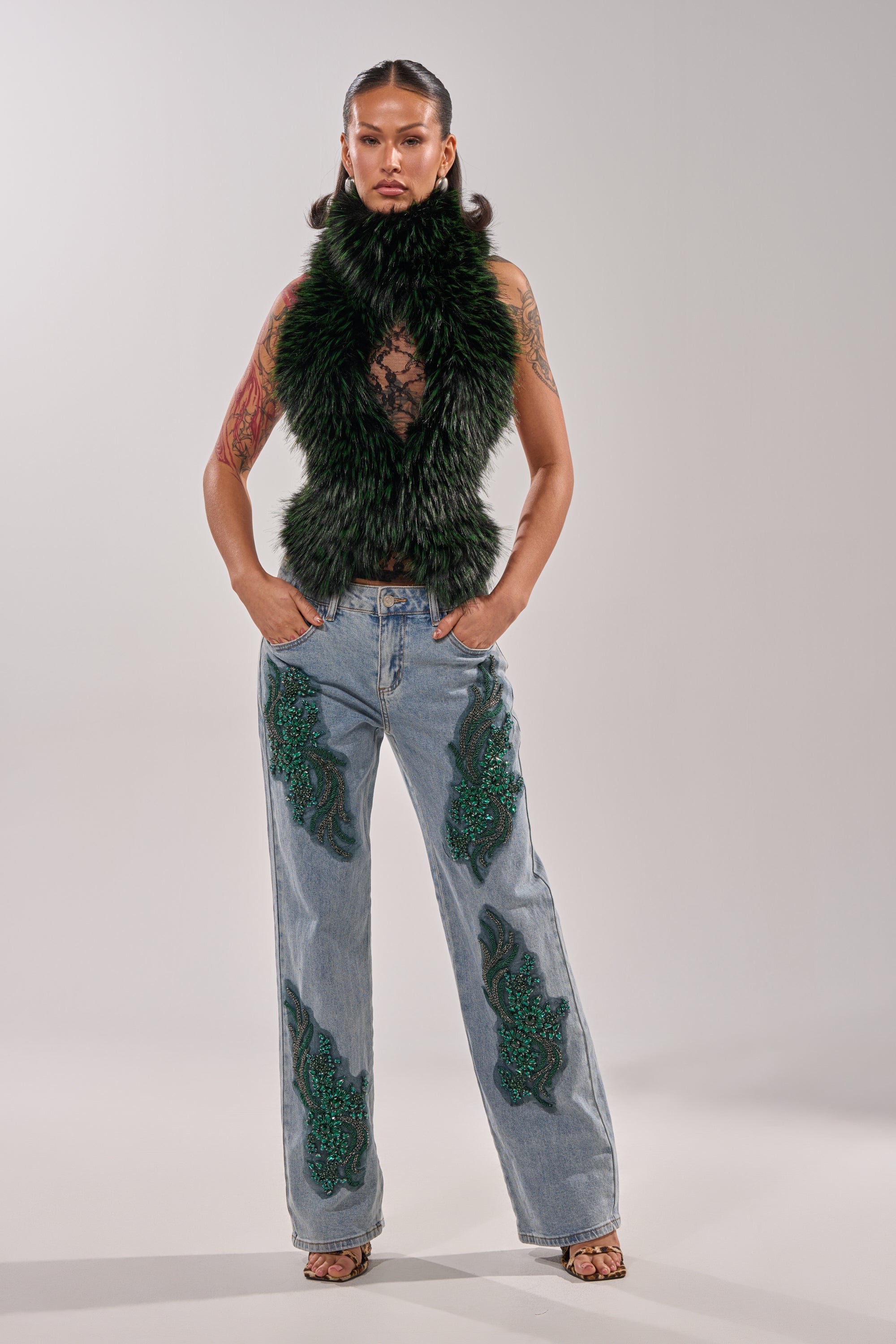 A woman poses confidently in BILLIONAIRE BELLE RHINESTONE JEANS, paired with a sleeveless dark green faux fur vest, sheer top, and open-toe heels against a plain gray backdrop. 1
