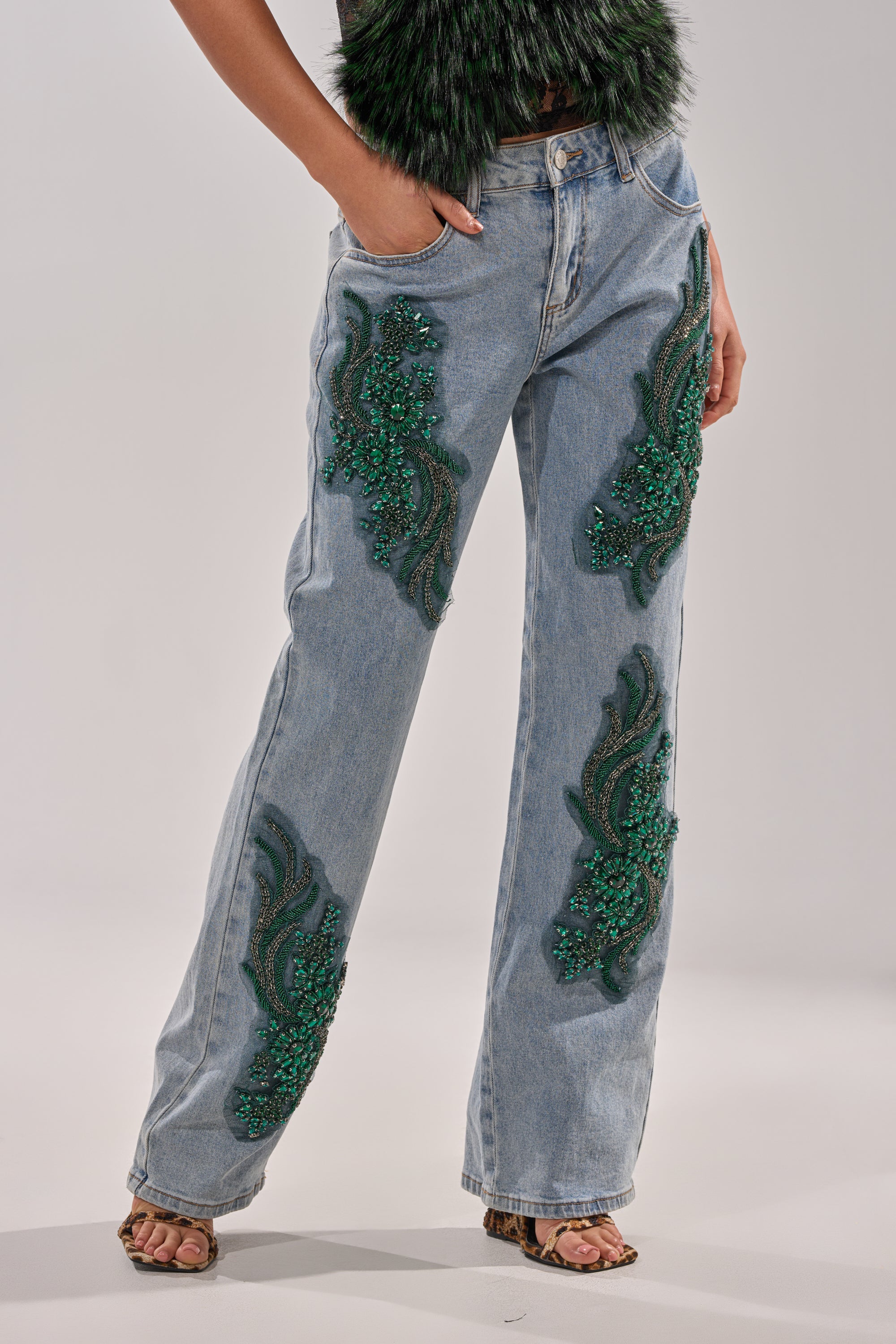 A person wears BILLIONAIRE BELLE RHINESTONE JEANS decorated with emerald beadwork on the thighs and lower legs, styled with a dark textured top and open-toe heels, set against a neutral, plain background. 0