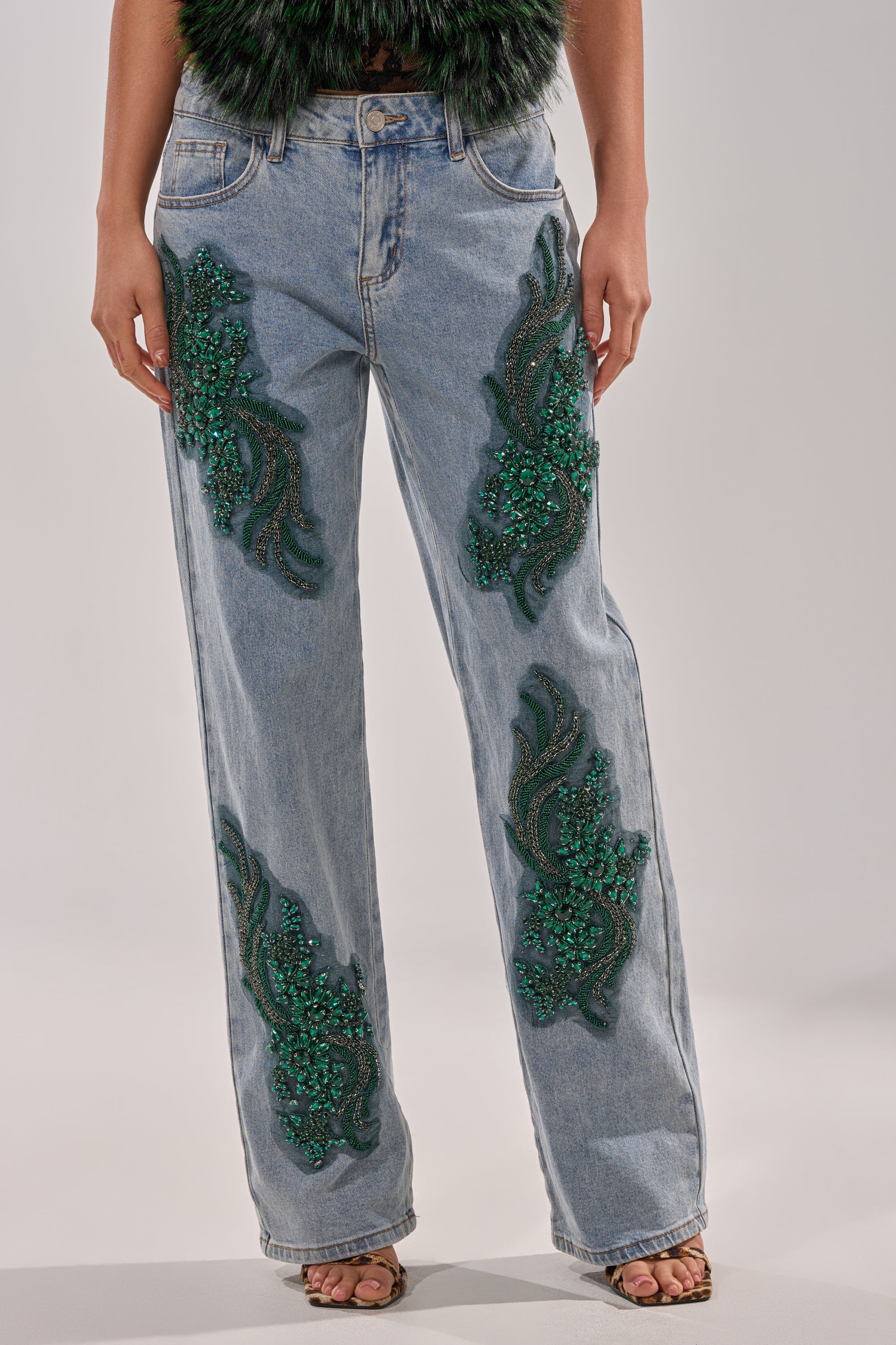 A person wears the BILLIONAIRE BELLE RHINESTONE JEANS—light blue, high-rise, wide-leg denim with emerald rhinestones and floral embroidery on the front—paired with open-toe heels. Their shirt is not visible in the image. 2