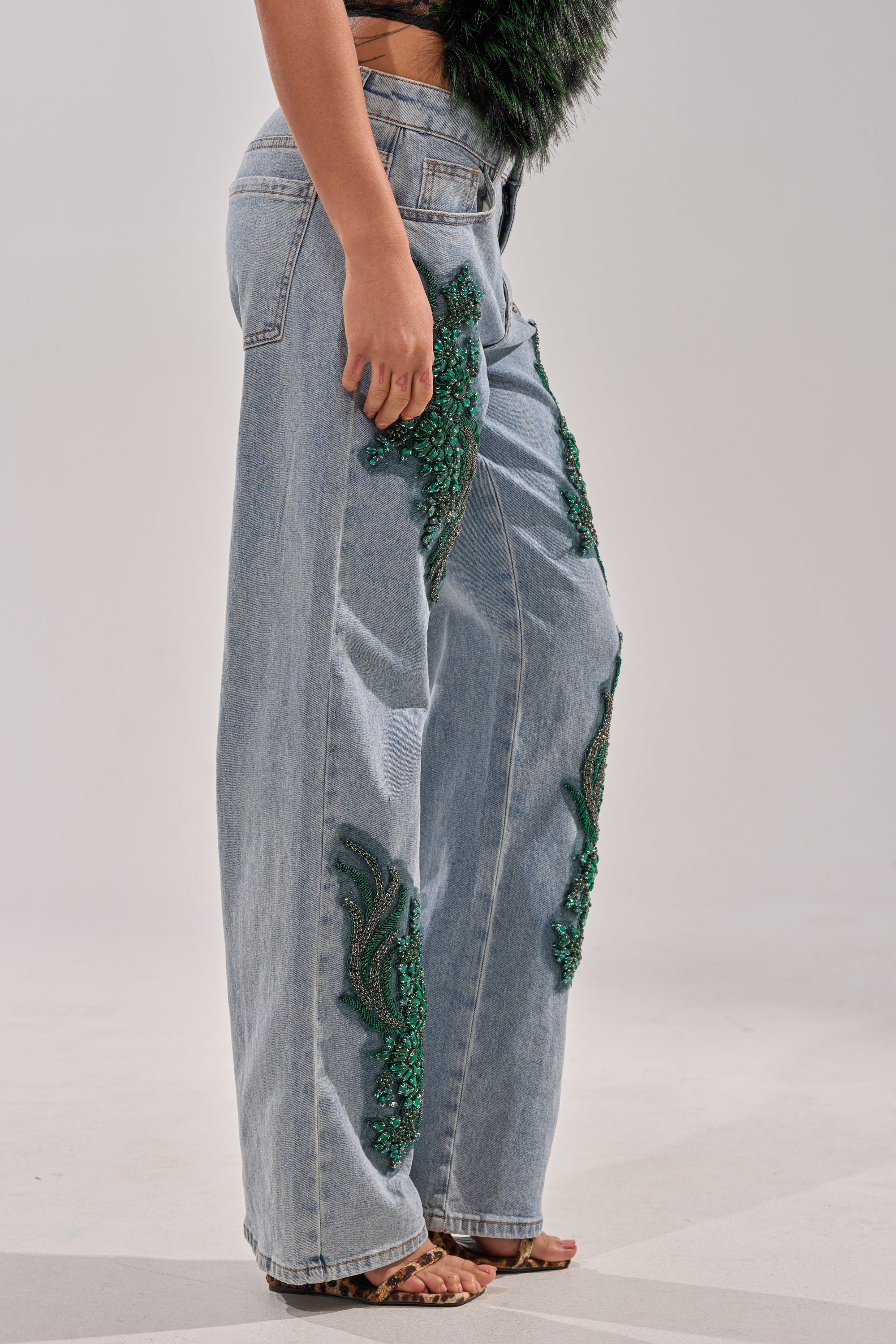 Someone wears the BILLIONAIRE BELLE RHINESTONE JEANS—light-wash, high-rise, with green rhinestone embellishments along the sides—paired with leopard print heeled sandals, standing against a plain light background. 3