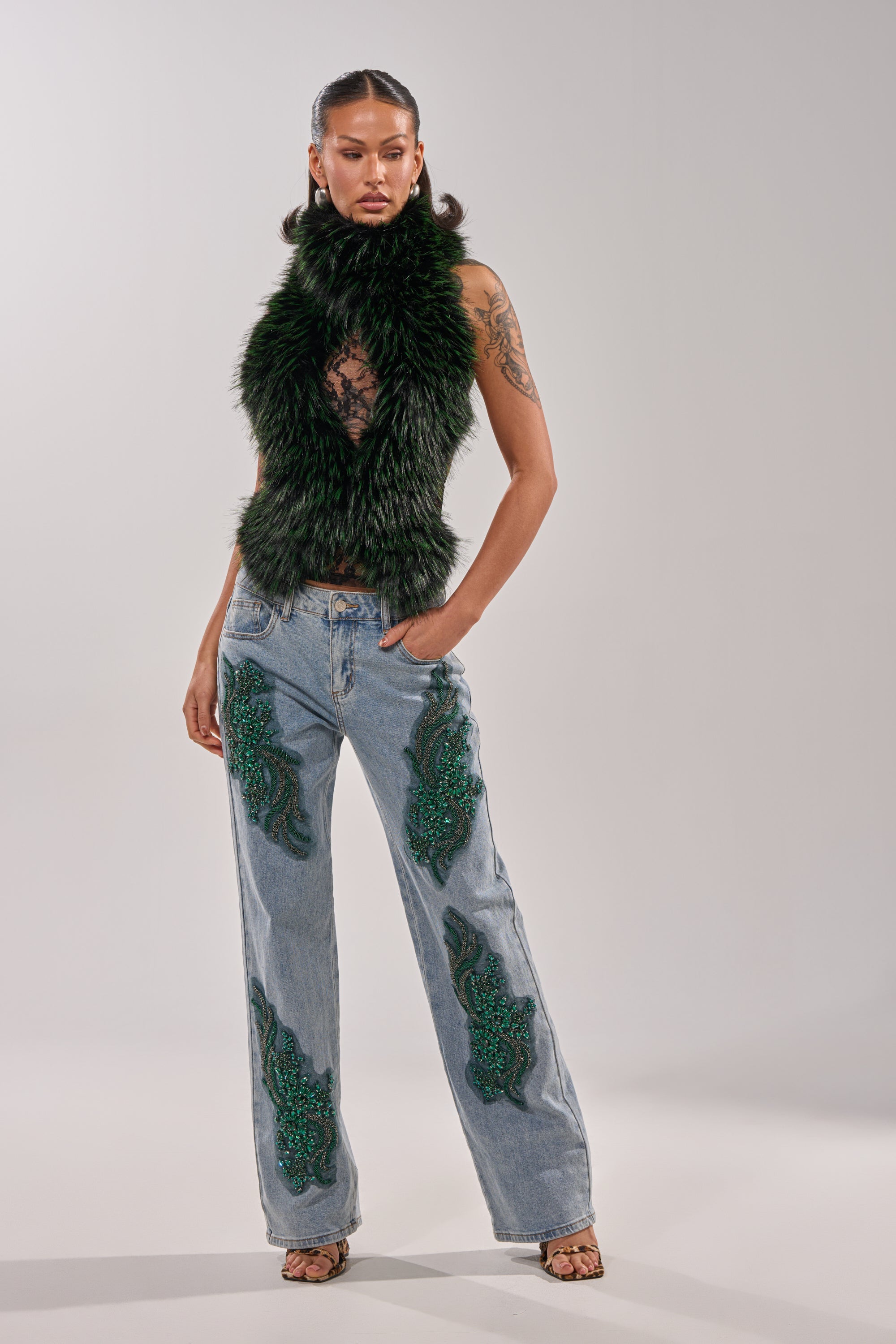A woman stands confidently in a black faux fur vest, sheer lace top, and the high-rise BILLIONAIRE BELLE RHINESTONE JEANS embellished with light-wash denim and green sequins. She completes the look with open-toe heels, arm tattoos, and pulled-back hair. 5