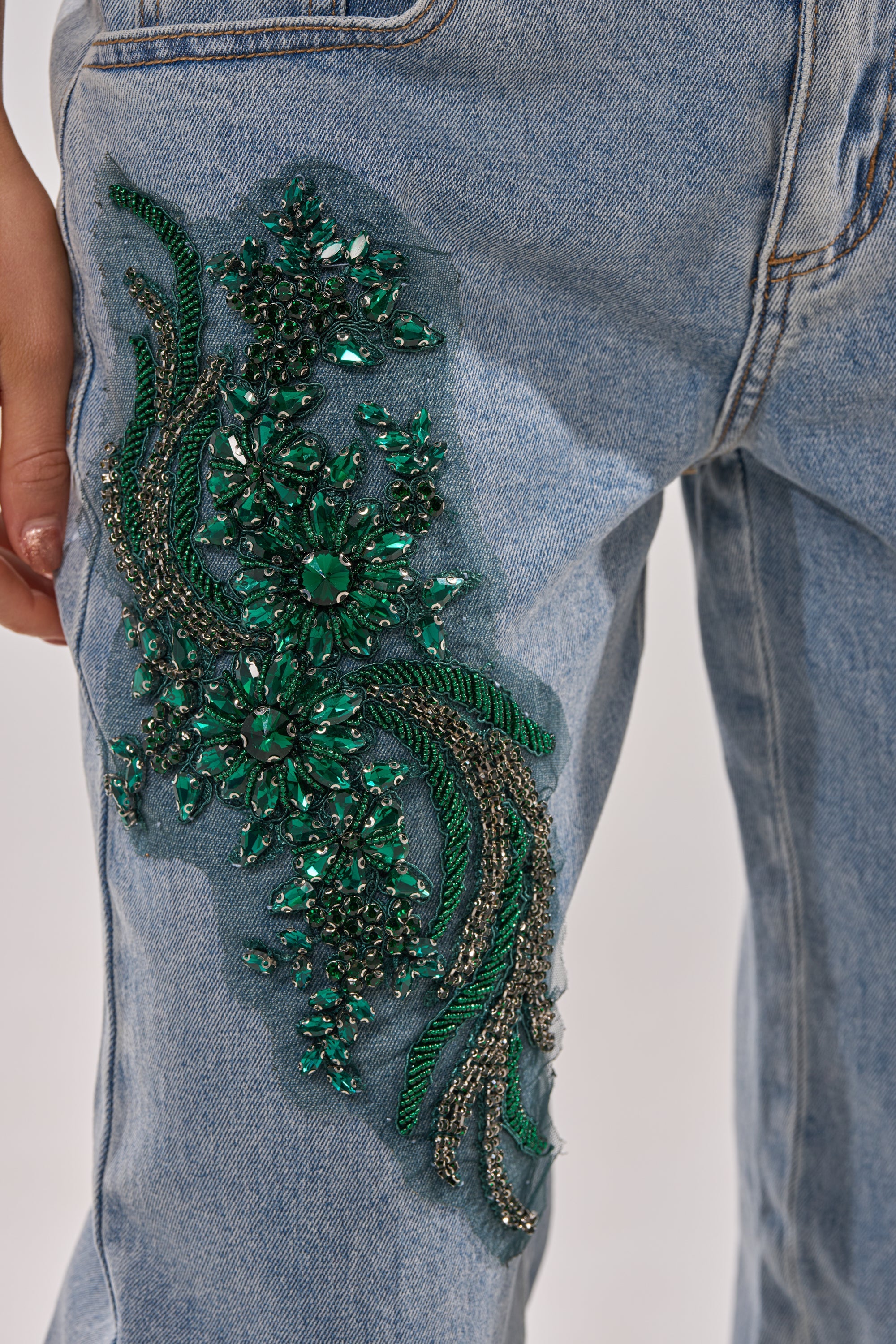 Close-up of BILLIONAIRE BELLE RHINESTONE JEANS in light-wash denim, showcasing a detailed green and silver beaded floral design on the upper thigh, with a hand resting nearby. 6