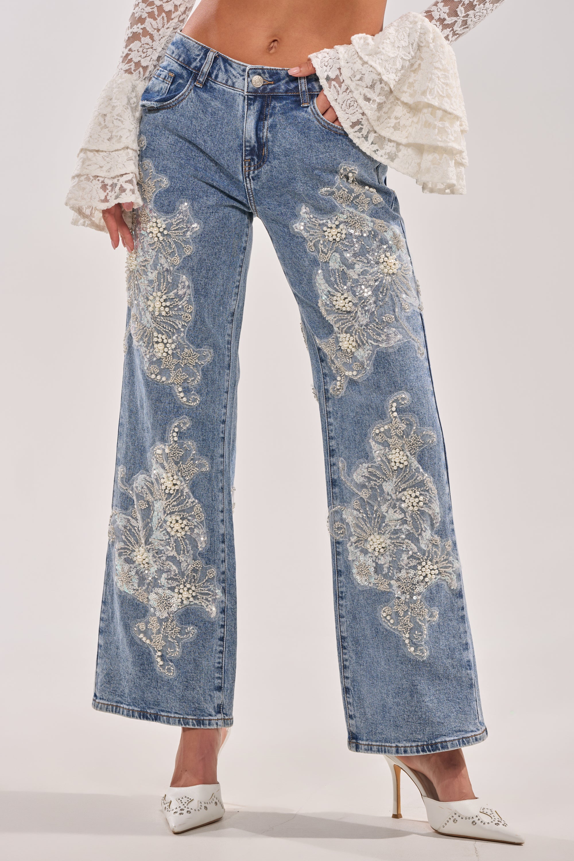 Wearing BILLIONAIRE BELLE RHINESTONE AND PEARL JEANS with intricate white lace and sequin floral embroidery, paired with a white lace ruffled-sleeve top and white pointed-toe heels.