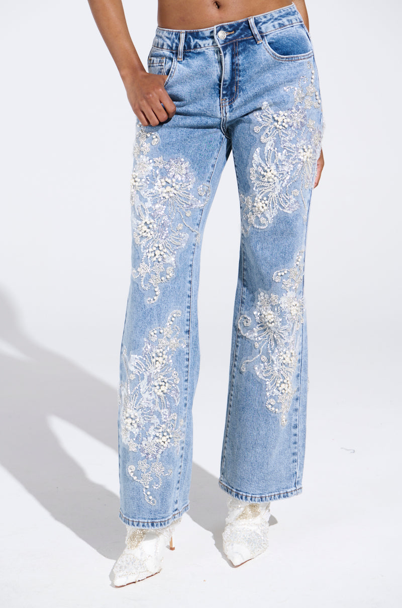 A person dons BILLIONAIRE BELLE RHINESTONE AND PEARL JEANS featuring intricate white floral embroidery and sequins with blue denim, paired with white pointed shoes. A plain white background highlights the jeans' detailed design.