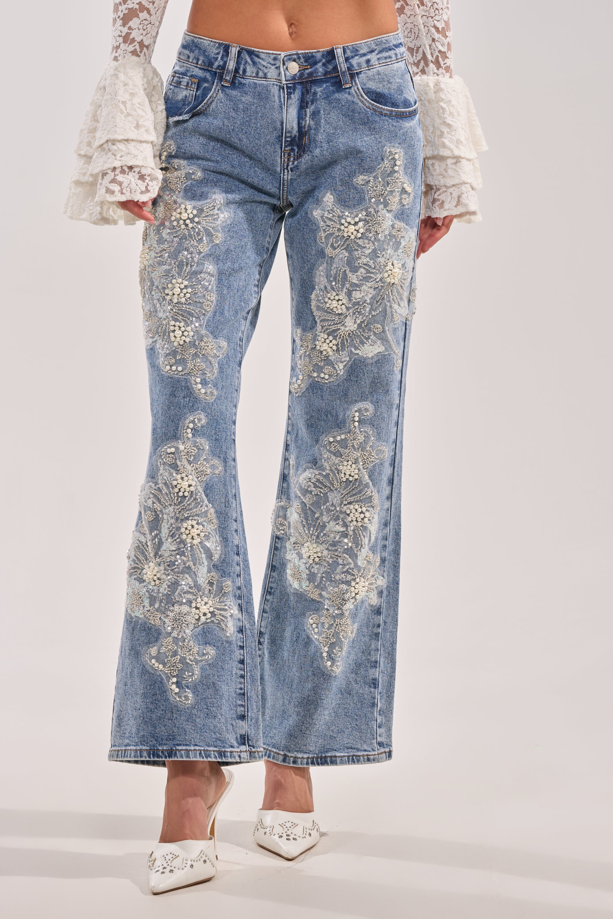 A person wearing BILLIONAIRE BELLE RHINESTONE AND PEARL JEANS, featuring luxury blue denim with rhinestone and pearl details, paired with a white lace ruffled-sleeve top and white pointed-toe shoes. 3
