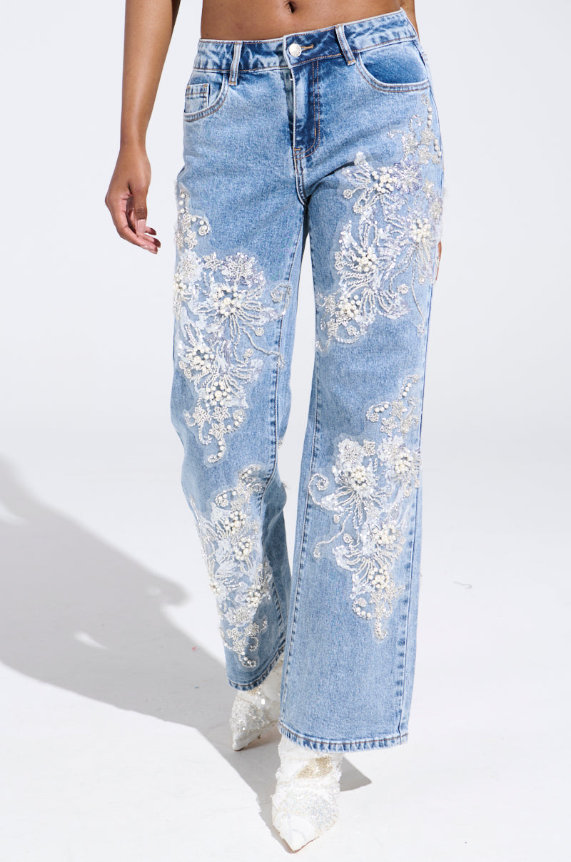 Someone wearing the striking BILLIONAIRE BELLE RHINESTONE AND PEARL JEANS featuring elaborate white floral embroidery on the legs, teamed with white pointed-toe shoes. The photo's simple white background accentuates the attire's elegance.