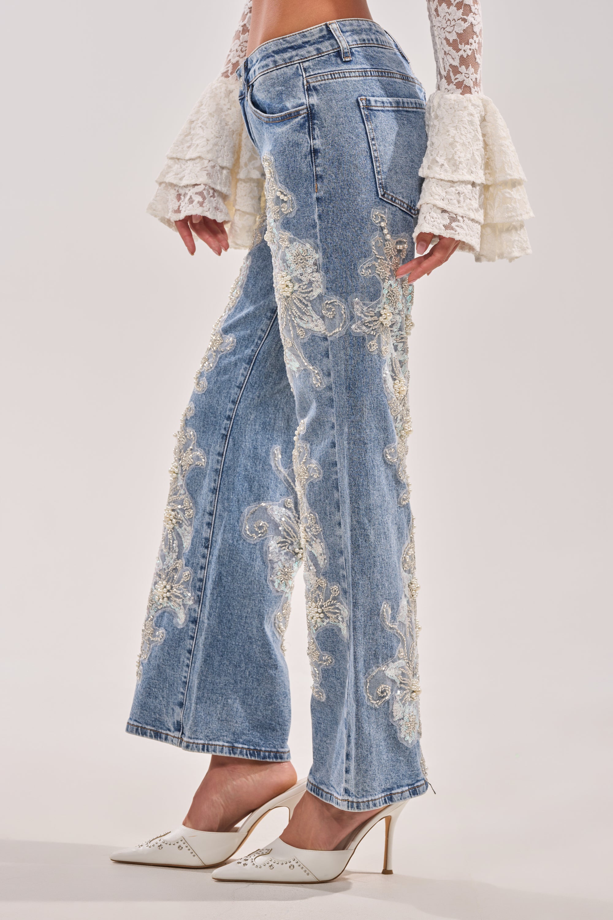 A person wearing BILLIONAIRE BELLE RHINESTONE AND PEARL JEANS, a white lace blouse with tiered ruffle sleeves, and white pointed-toe heels. Only the lower half of the body is visible. 4