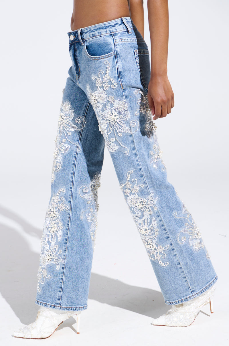A side view of legs adorned in light blue rhinestone and pearl jeans with intricate white lace embroidery, paired with chic white high heels, elegantly showcases the Billionaire Belle against a plain backdrop.