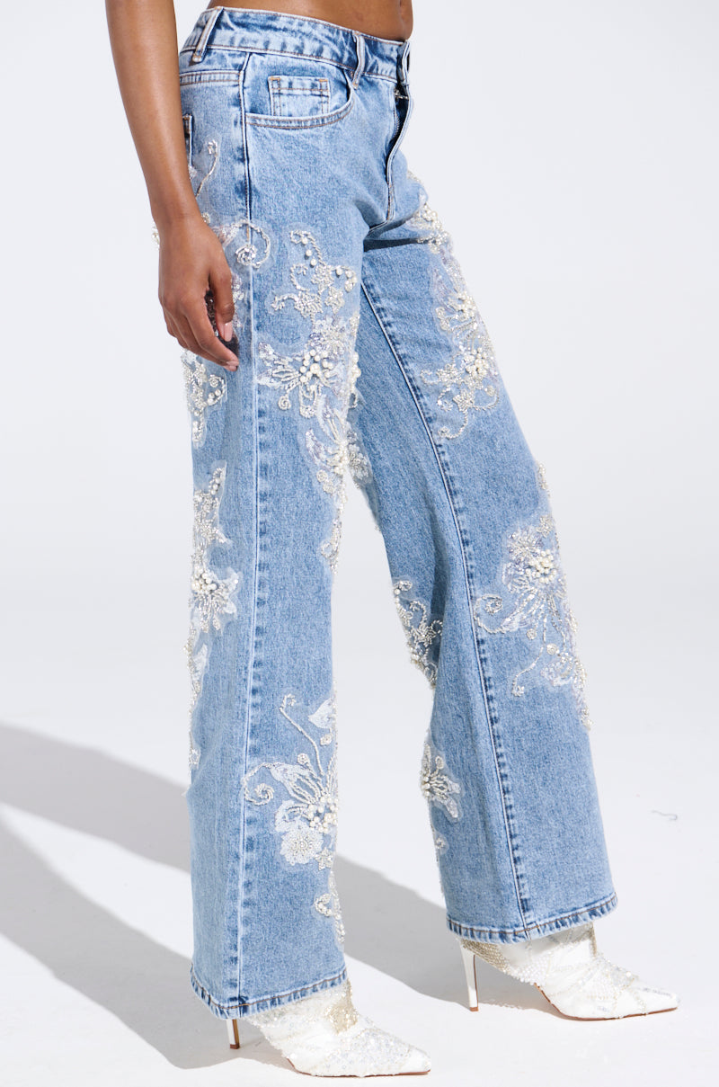 Dazzling in blue high-waisted, wide-leg jeans from the Billionaire Belle collection, adorned with rhinestones and white floral lace embroidery, paired with white pointed-toe lace-textured ankle boots. A simple light backdrop enhances the chic Billionaire Belle vibe.