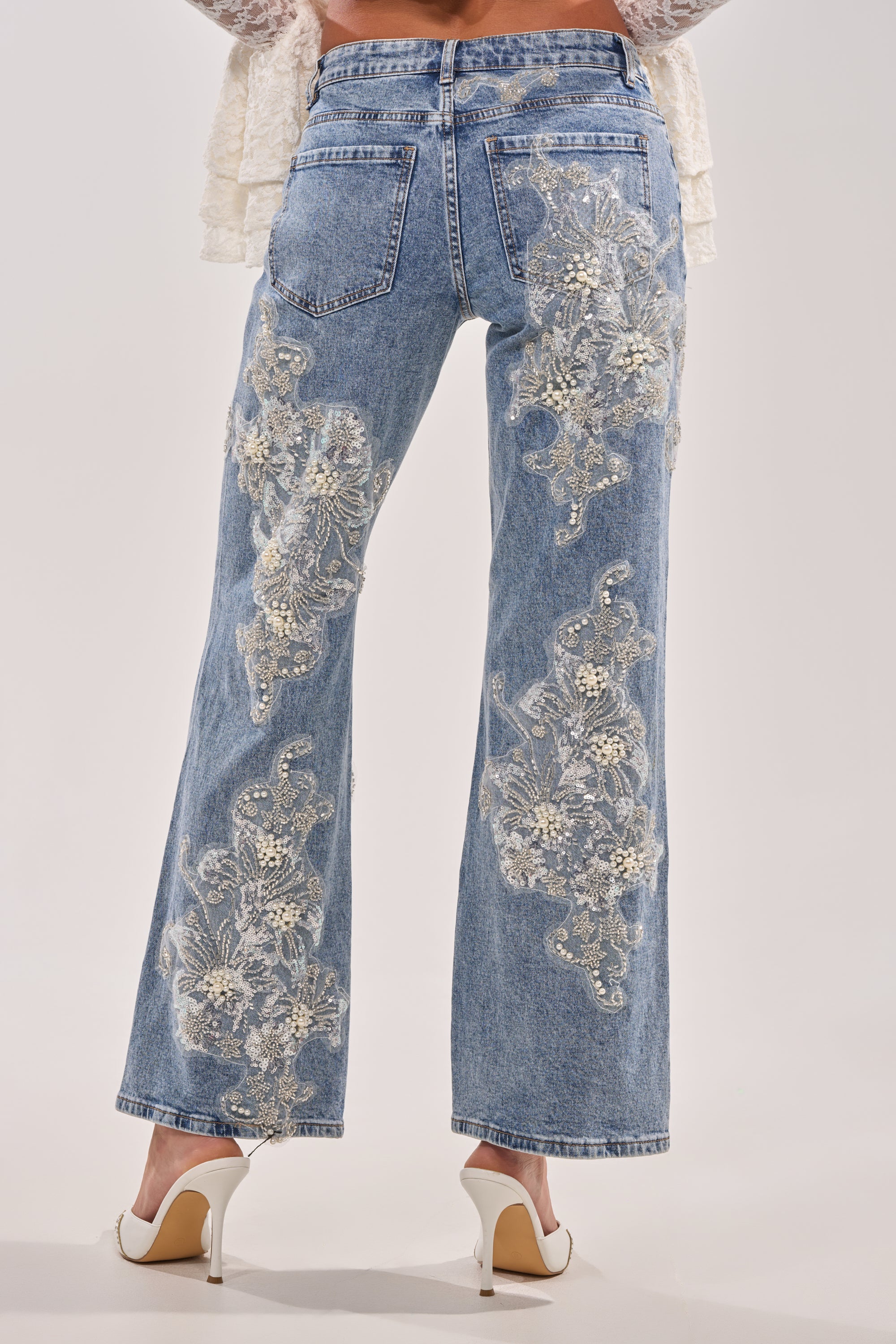 A person models the BILLIONAIRE BELLE RHINESTONE AND PEARL JEANS, luxury high-waisted denim adorned with intricate rhinestone and pearl embellishments on the back, paired with white heels against a plain background. 6