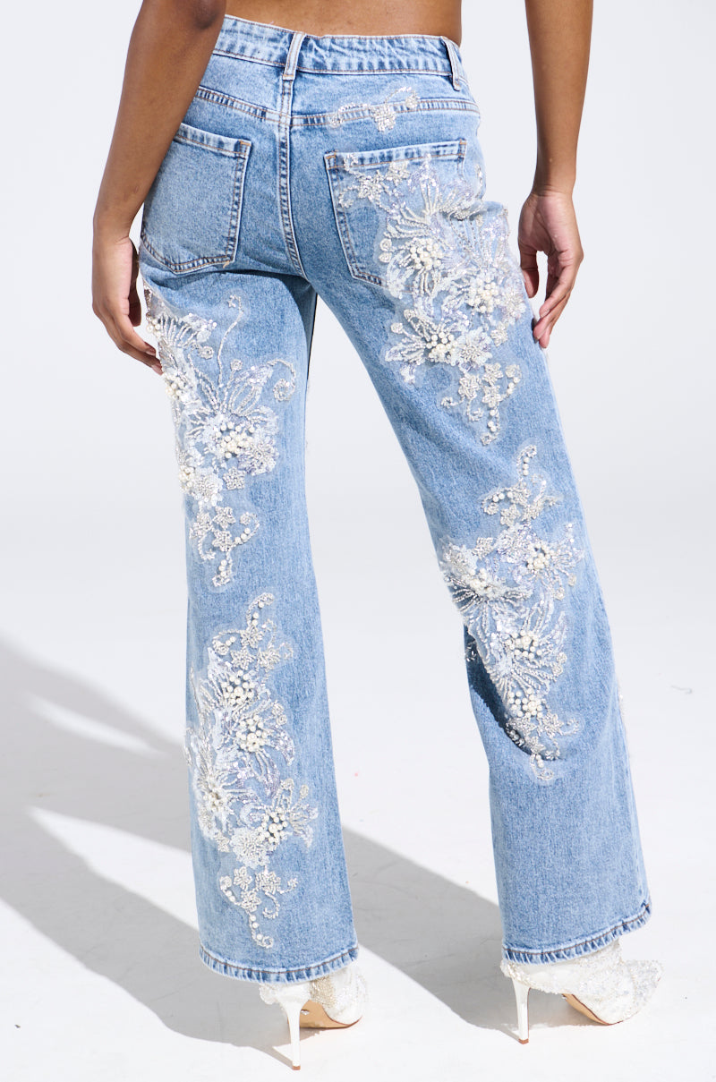 A person wearing BILLIONAIRE BELLE RHINESTONE AND PEARL JEANS, adorned with white floral lace embroidery on light blue denim, exuding elegance. The high-waisted jeans with rear pockets pair perfectly with chic white open-toed heels.