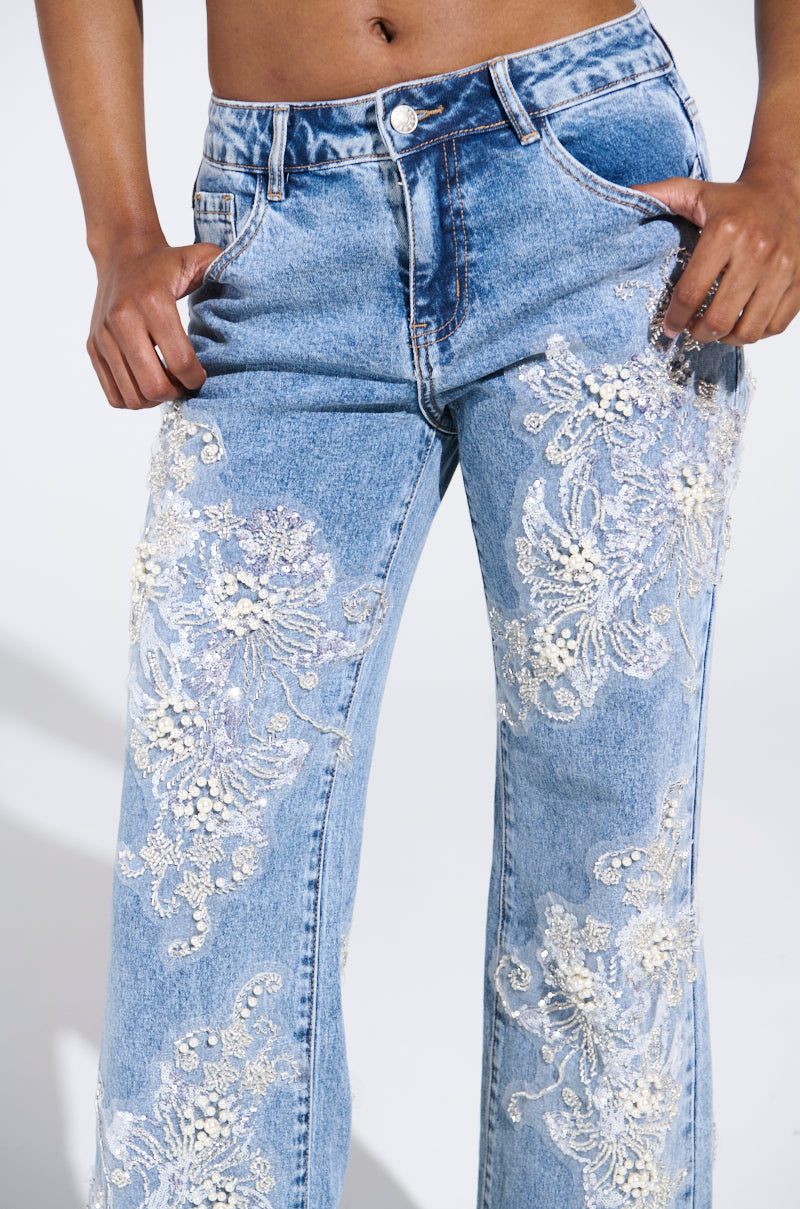 A close-up showcases the BILLIONAIRE BELLE RHINESTONE AND PEARL JEANS, with white lace floral embroidery and rhinestone accents. The light blue denim features front pockets and a button closure, as hands rest confidently on hips against a plain white backdrop.