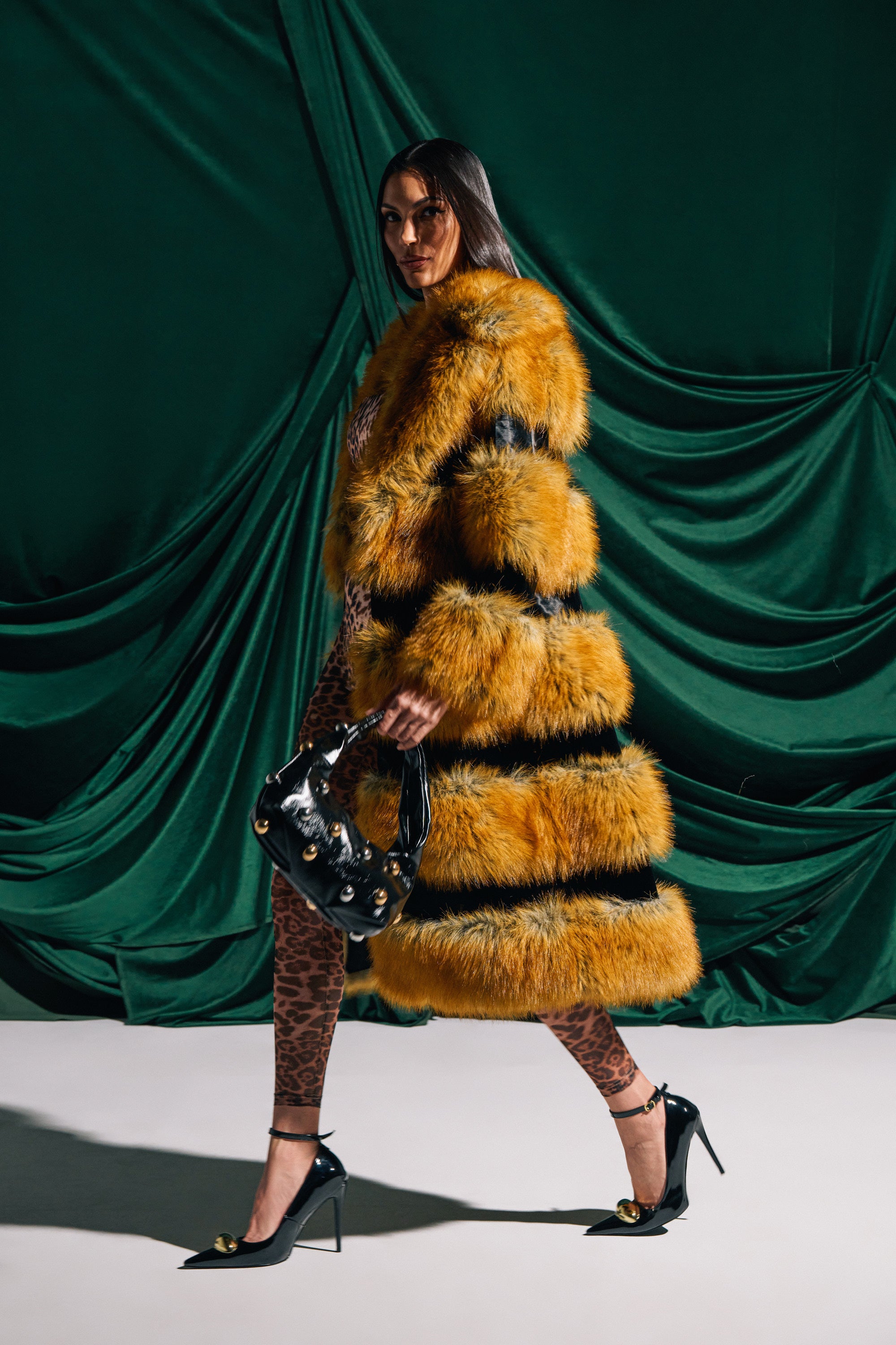 A woman in the bold MADDOX PANELED FAUX FUR coat and leopard print pants walks confidently with a black handbag and black heels with gold accents, set against a dark green velvet backdrop.
