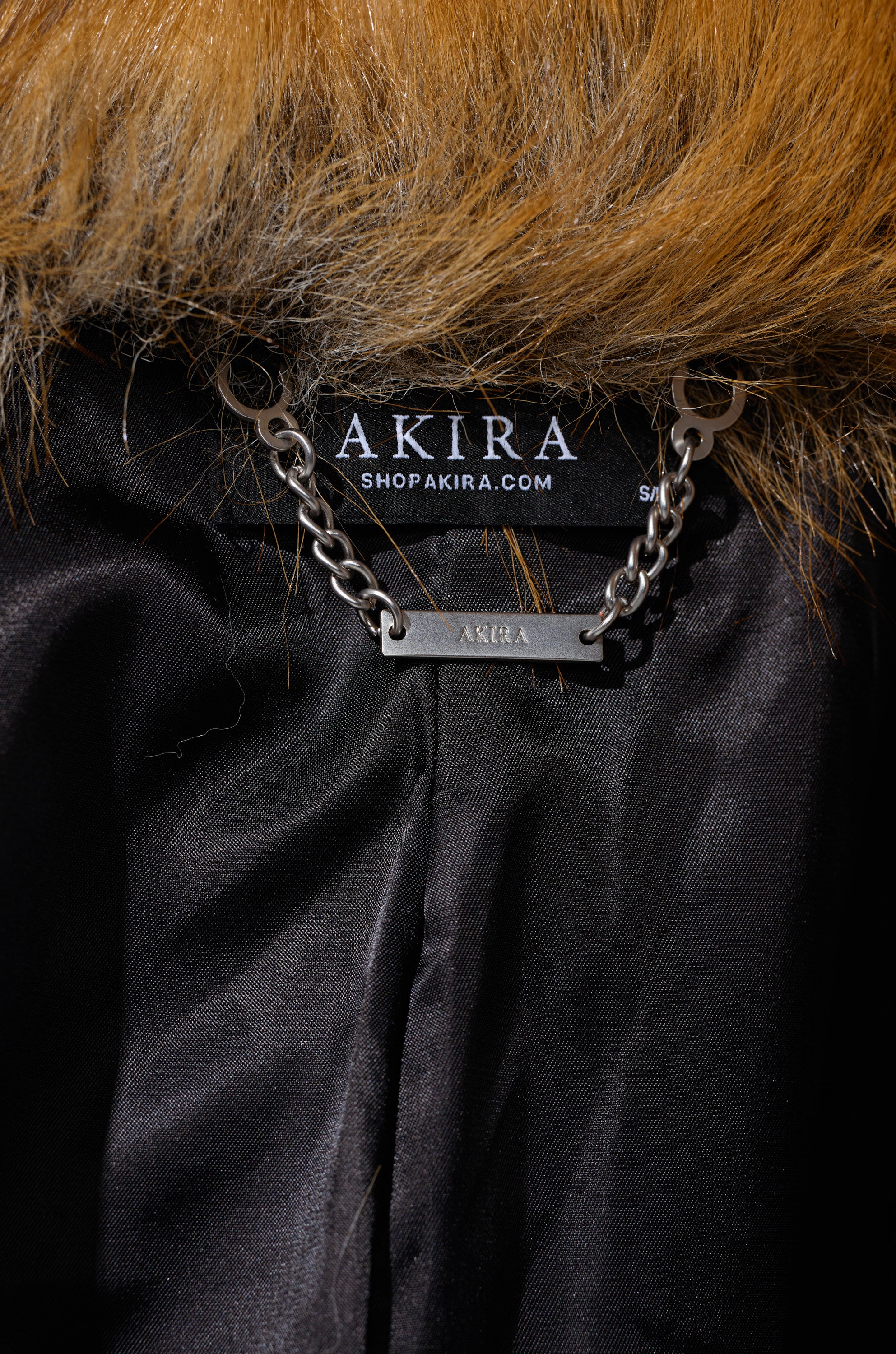 Close-up of the MADDOX PANELED FAUX FUR: a black longline coat with a brown faux fur collar, AKIRA label, shopakira.com tag, and a metal chain nameplate.