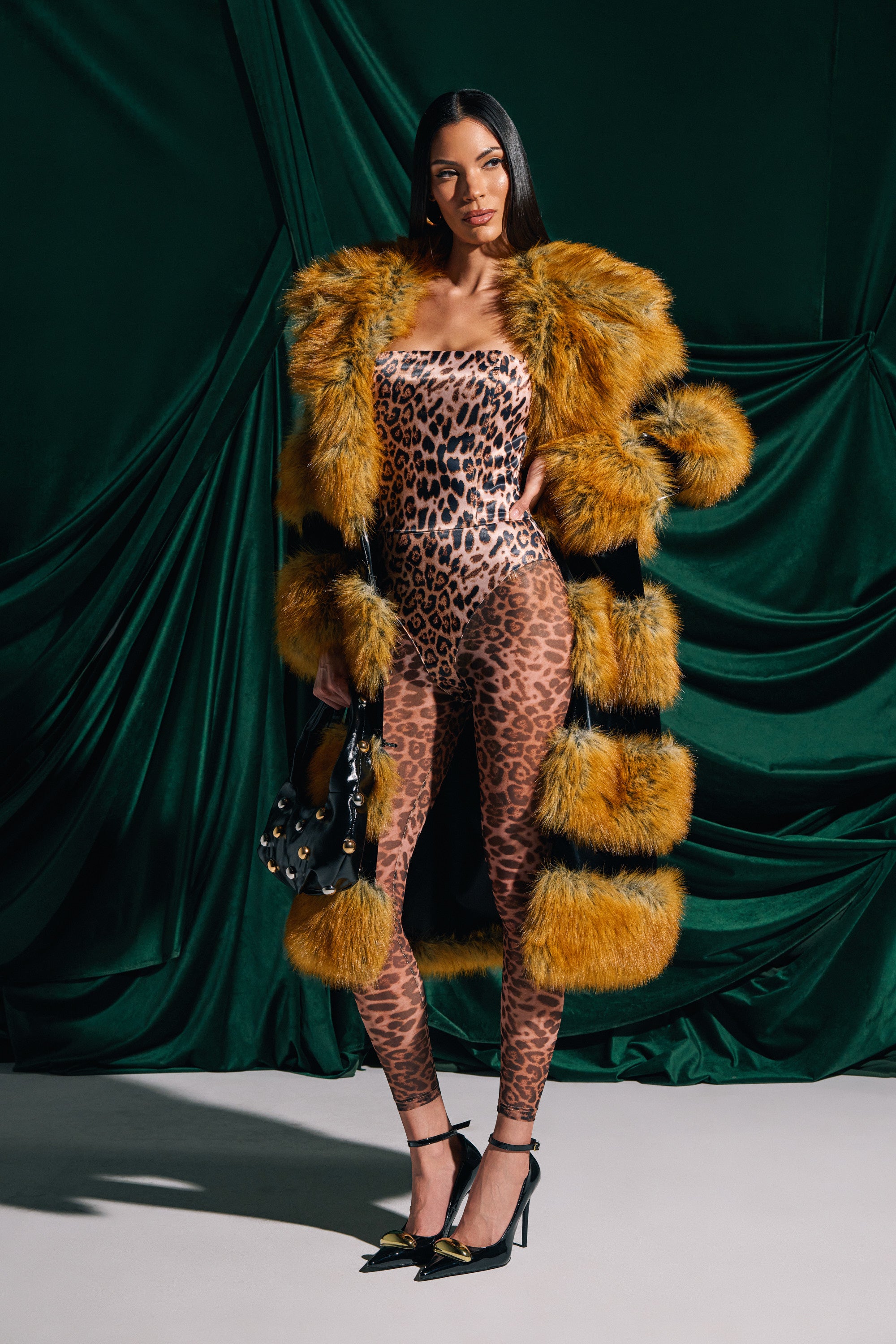 A woman stands confidently against a dark green draped backdrop, wearing the bold MADDOX PANELED FAUX FUR coat with orange and black stripes and an oversized collar over a sleek leopard print bodysuit. She accessorizes with a studded handbag and high heels.