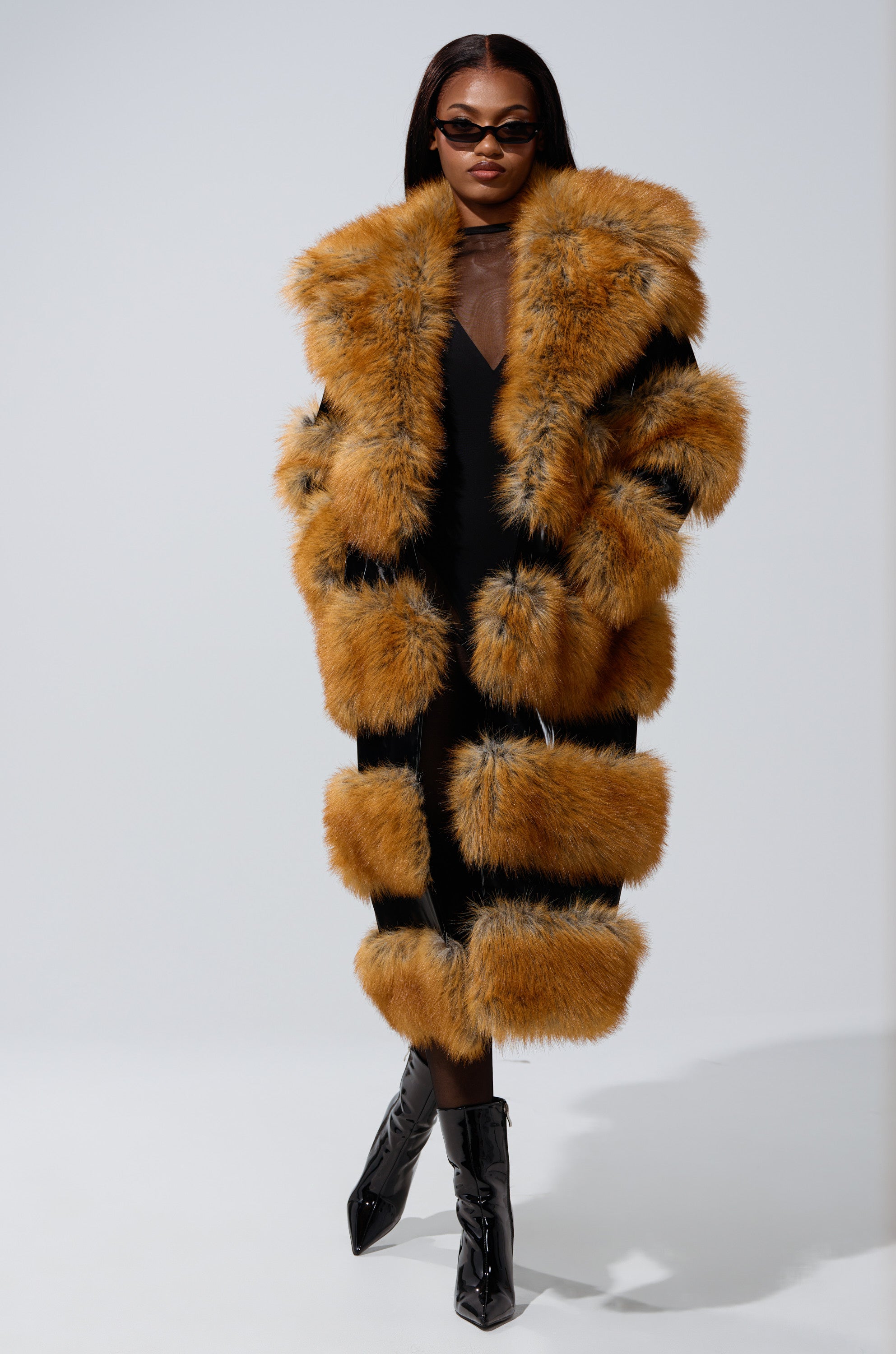 Wearing dark sunglasses, a long black dress, shiny black ankle boots, and the MADDOX PANELED FAUX FUR coat with an oversized fur collar, a person stands confidently against a light background.