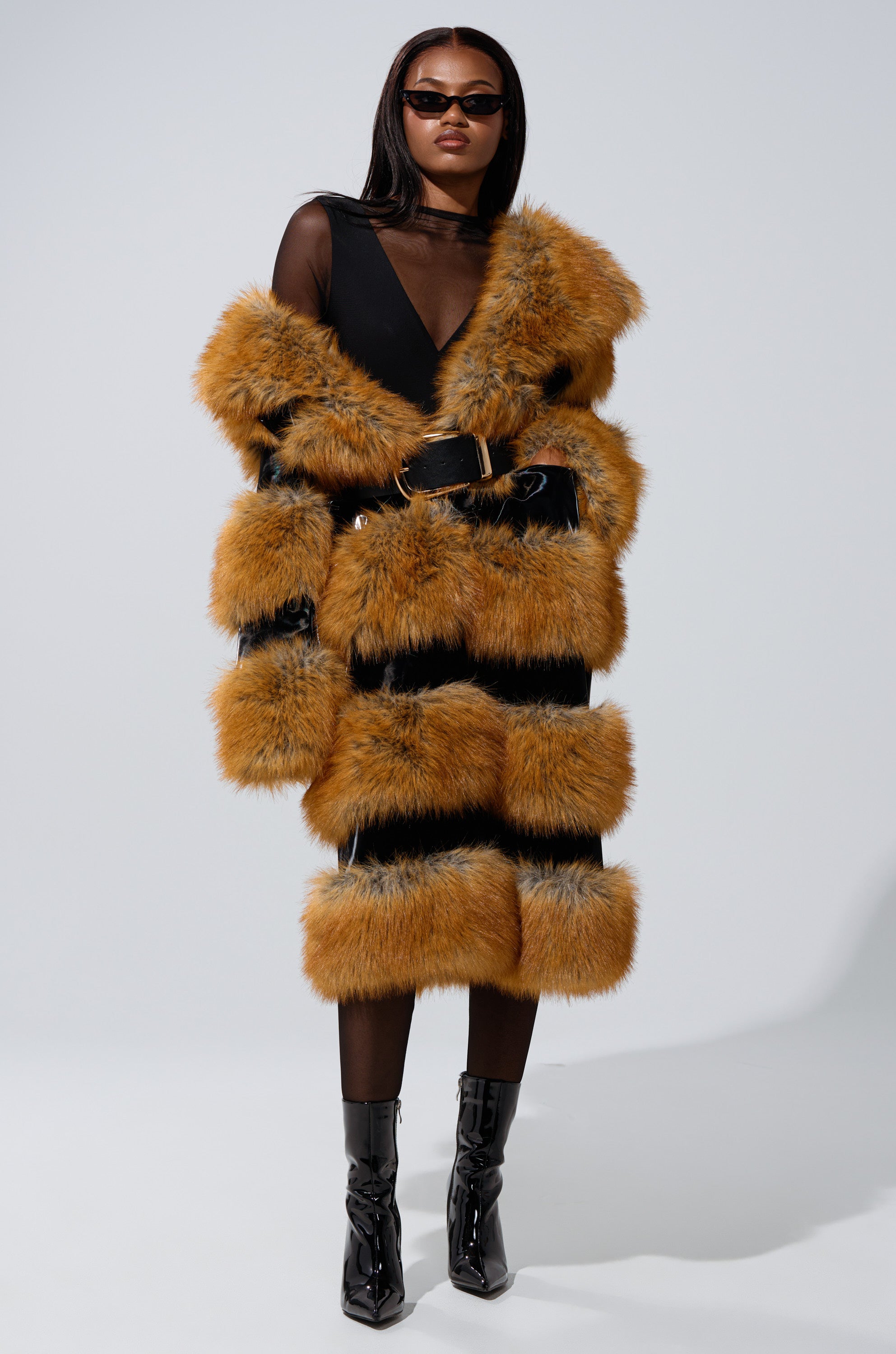 A woman stands against a plain light background wearing sunglasses, a black sheer top, and the MADDOX PANELED FAUX FUR coat in striped brown and black, styled with a black belt and shiny black ankle boots.