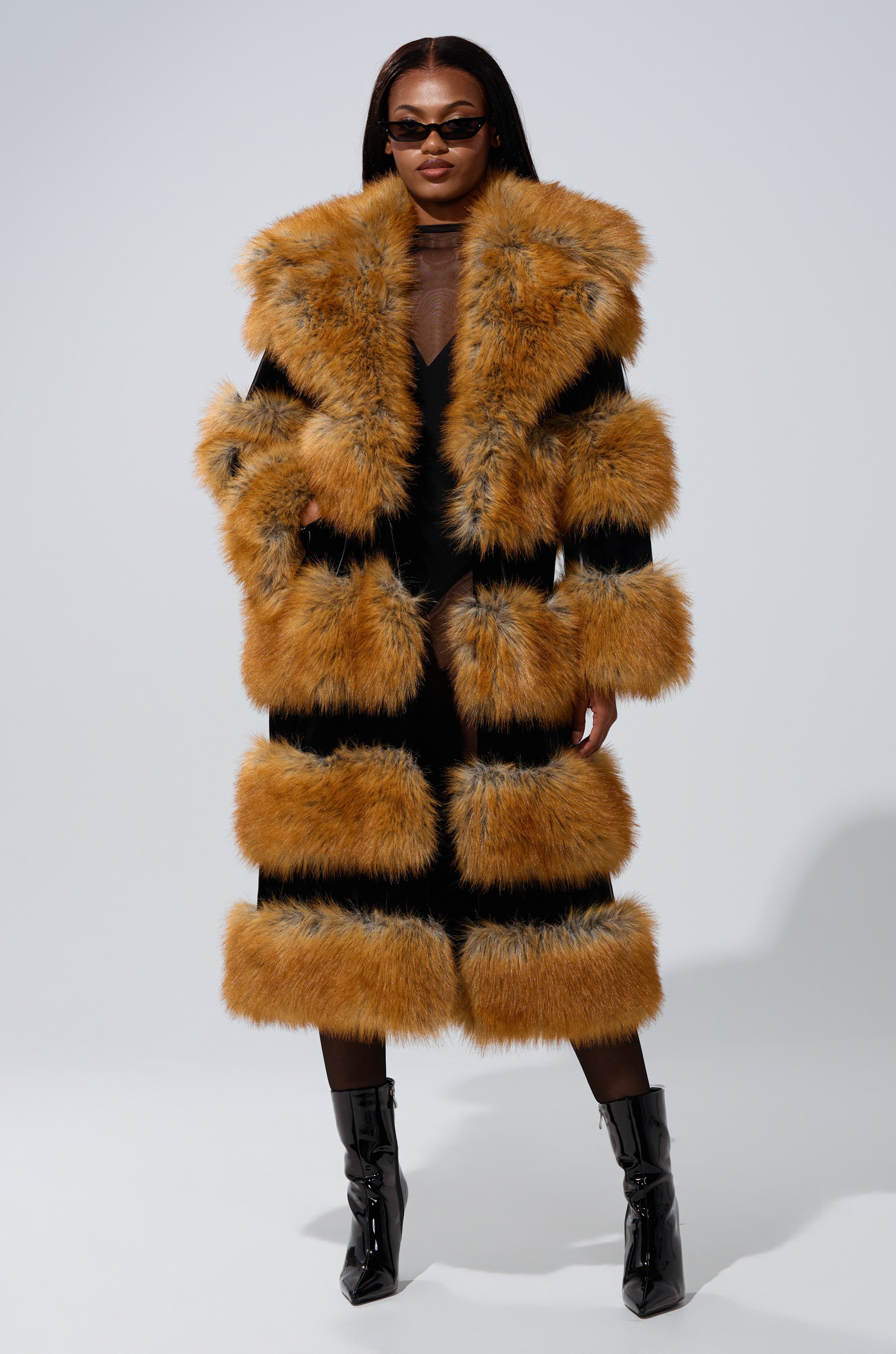 A woman exudes confidence in dark sunglasses and the MADDOX PANELED FAUX FUR coat, featuring bold horizontal orange-brown panels. She completes her striking look with black boots against a plain gray background.
