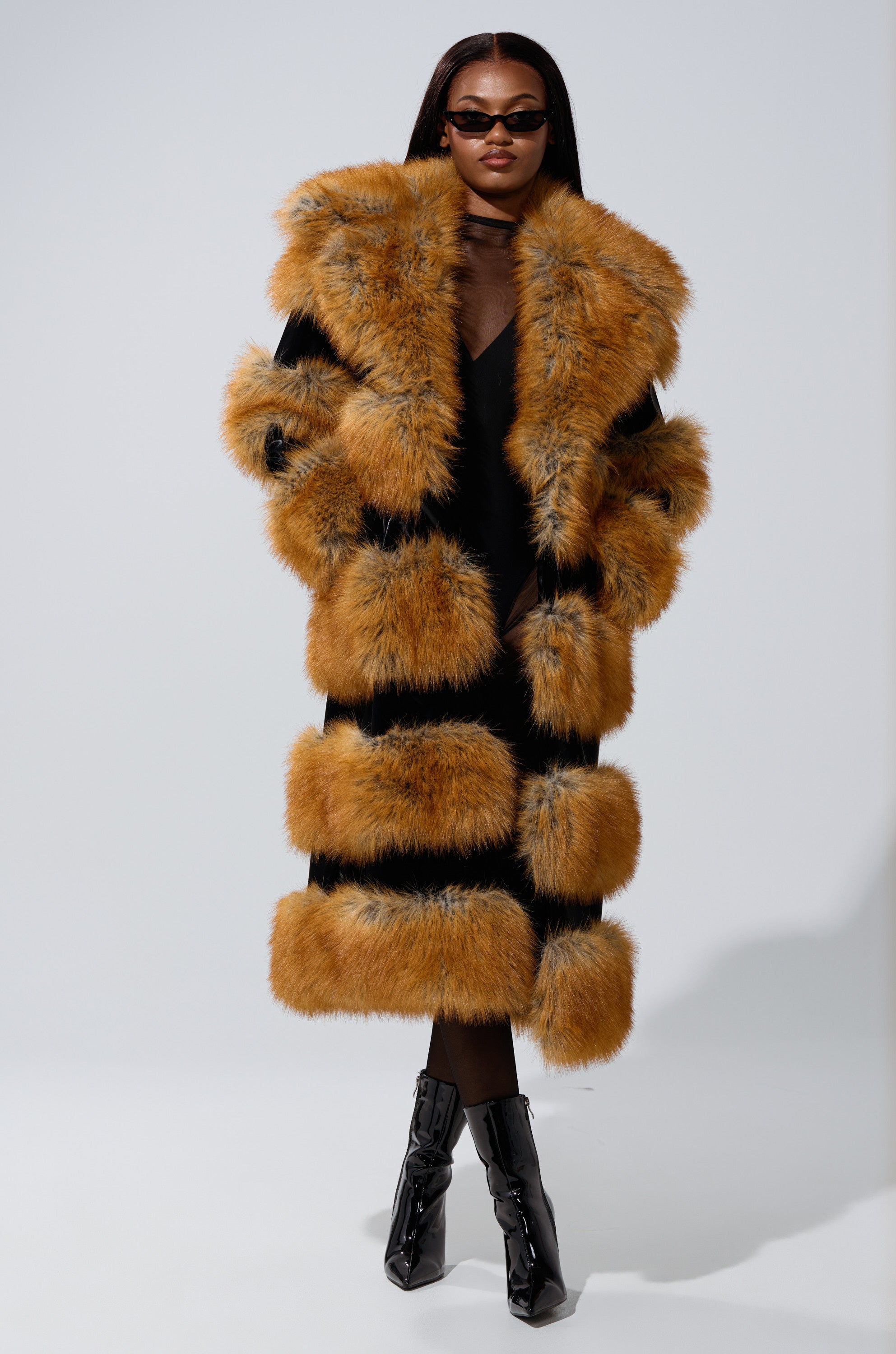 A woman wearing the MADDOX PANELED FAUX FUR coat, black ankle boots, and dark sunglasses stands against a light gray background, highlighting bold statement outerwear.