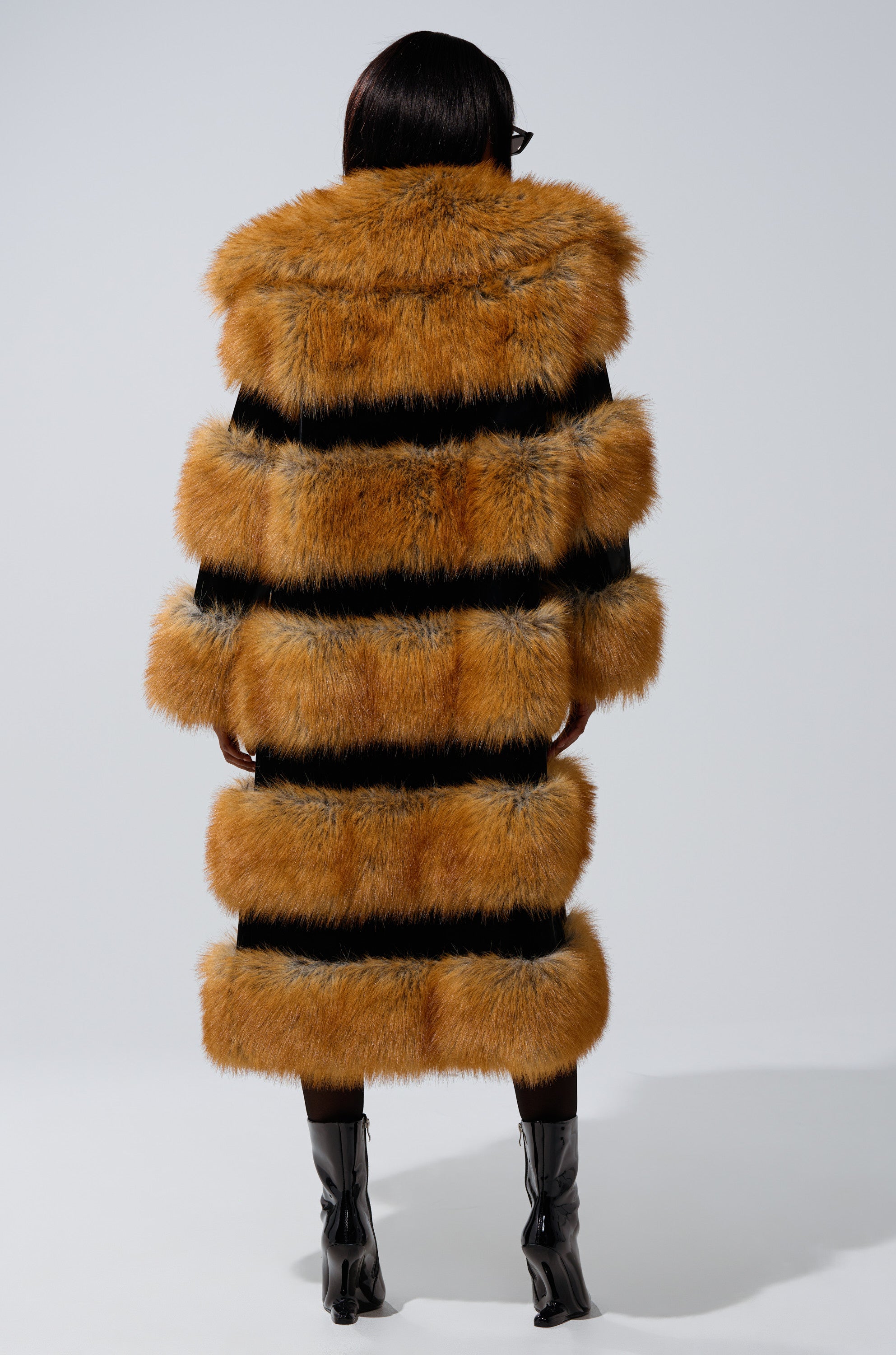 A person faces away, wearing the MADDOX PANELED FAUX FUR coat with orange and black stripes, an oversized fur collar, black boots, and dark sunglasses against a plain light background.