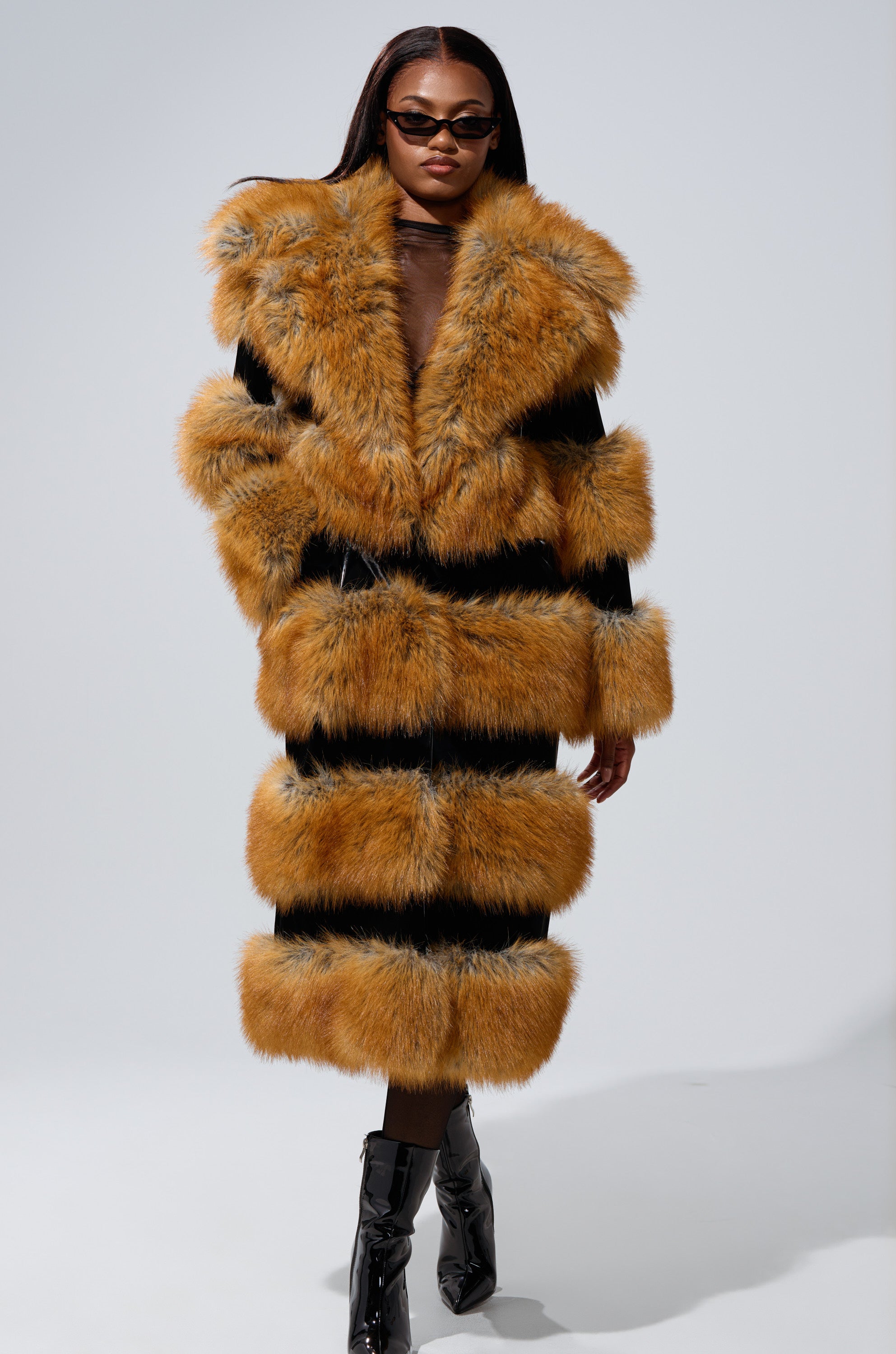 Wearing the MADDOX PANELED FAUX FUR coat with wide orange-brown and black panels, a woman in dark sunglasses pairs it with black ankle boots as she poses confidently against a plain light background.