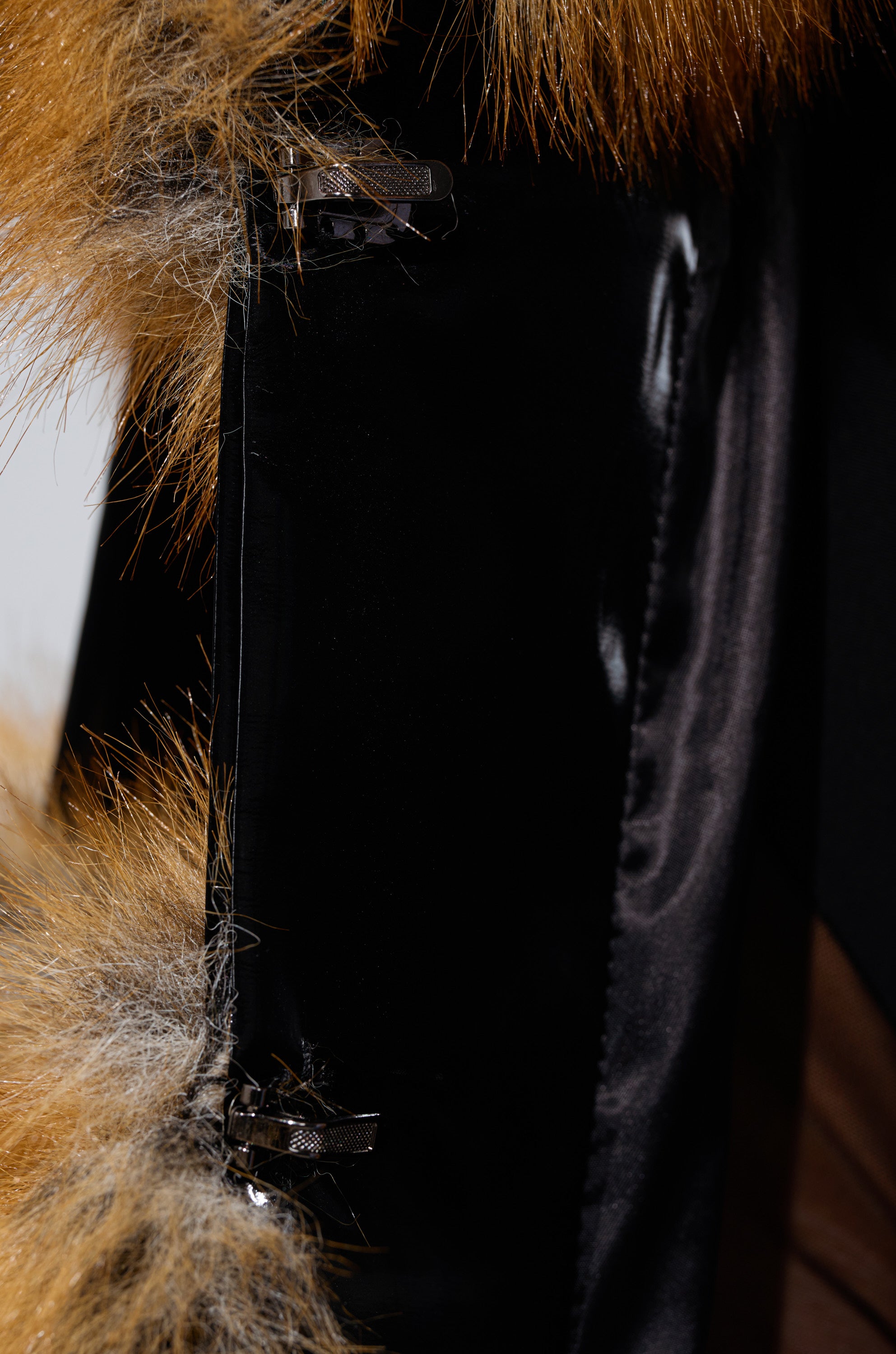 Close-up of the MADDOX PANELED FAUX FUR coat, featuring an oversized brown faux fur collar with white streaks, glossy black satin lining, and two metal hook-and-eye clasps. The luxurious fabric appears smooth to the touch.