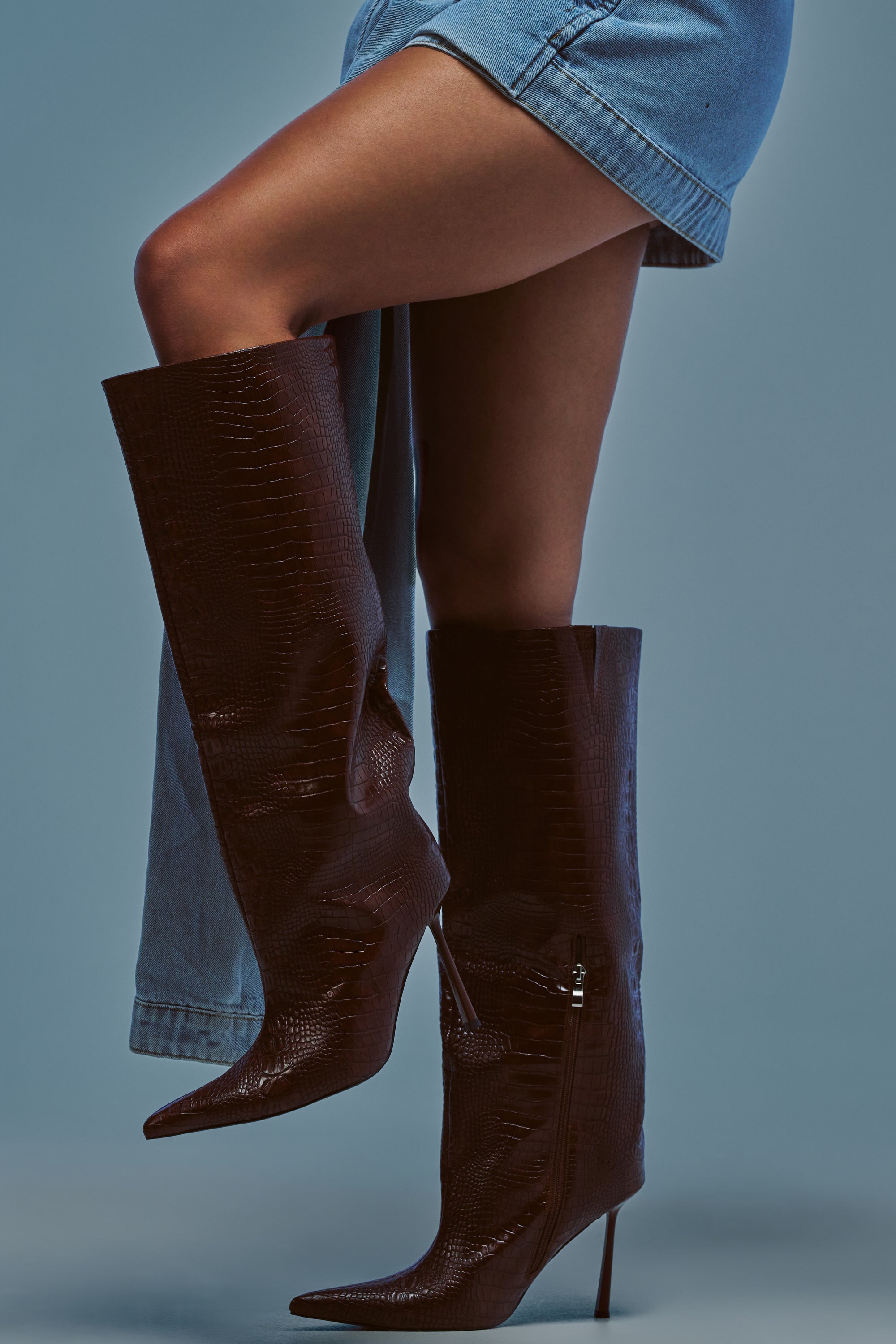 A person wearing a denim skirt and striking AZALEA WANG MADEYE BROWN CROC KNEE HIGH STILETTO BOOTS stands against a plain background.