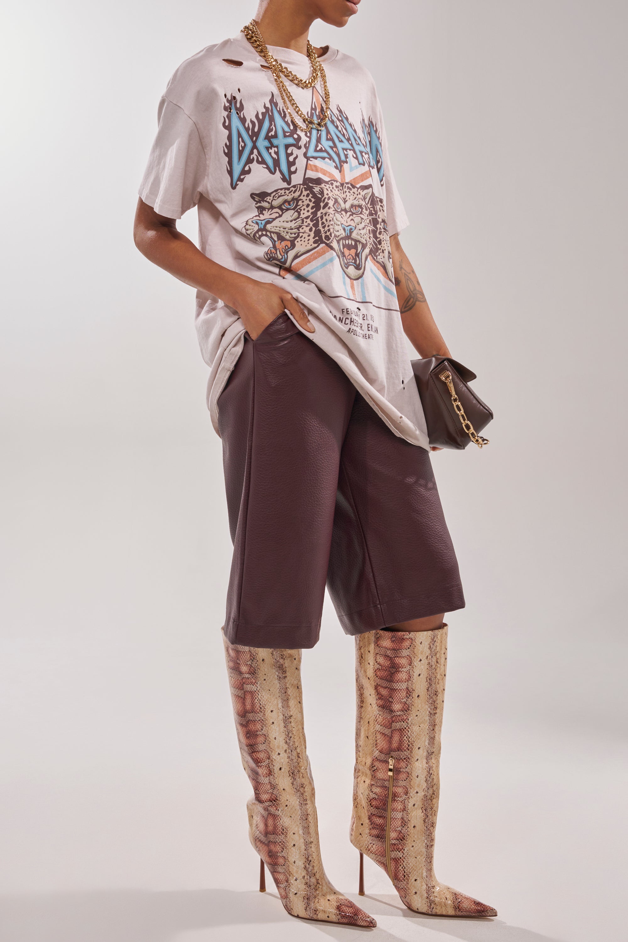 A person wears a distressed Def Leppard tee, dark loose shorts, layered gold necklaces, a small brown clutch, and AZALEA WANG MADEYE NUDE SNAKE STILETTO BOOTS, creating a stylish and edgy look. 4