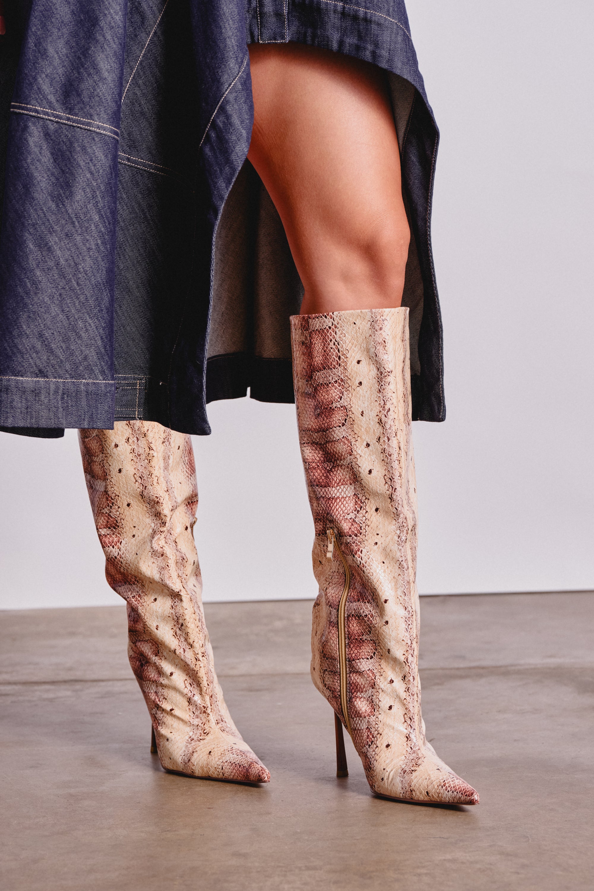 A person stands on a concrete floor in a dark denim skirt and AZALEA WANG MADEYE NUDE SNAKE STILETTO BOOT, featuring pointed toes and faux leather snakeskin knee-high design. 1