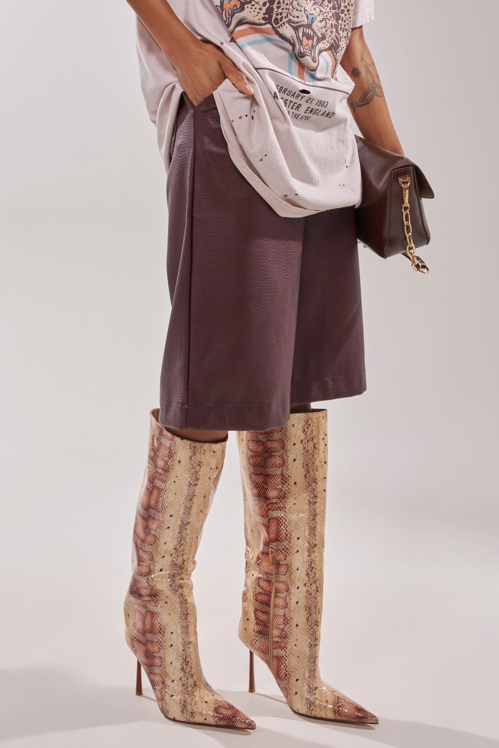 The lower half of a person shows them in loose brown knee-length shorts, a graphic white tee, and holding a brown clutch with a gold chain, wearing AZALEA WANG MADEYE NUDE SNAKE STILETTO BOOTS with pointed toes. 6