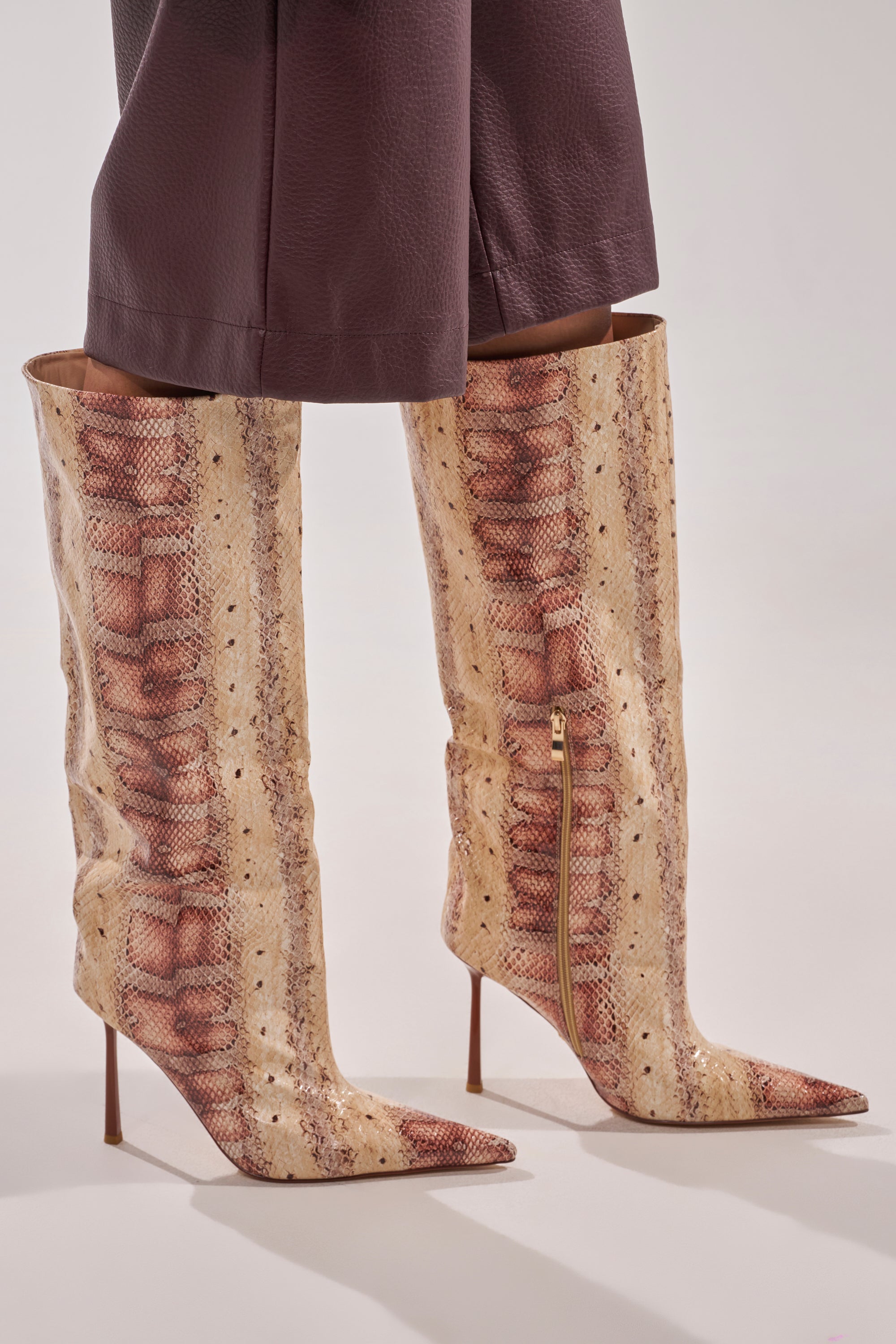 A person wears the AZALEA WANG MADEYE NUDE SNAKE STILETTO BOOT—knee-high, beige and brown snakeskin with pointed toes and side zippers—paired with loose, dark pants. 3