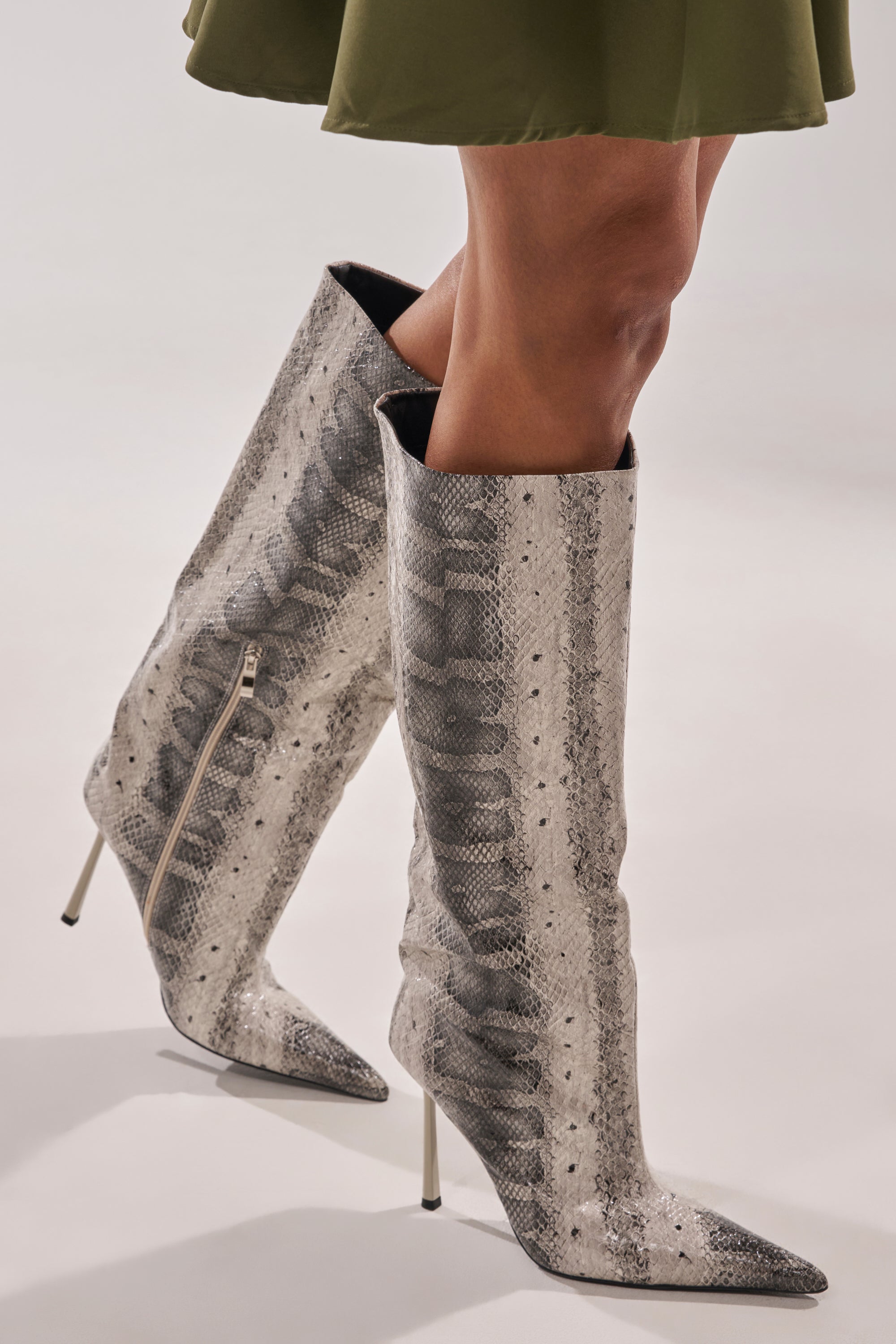 A person wearing the AZALEA WANG MADEYE SNAKE STILETTO BOOT—knee-high, pointed-toe boots in a gray and black snakeskin pattern—paired stylishly with an olive green skirt.