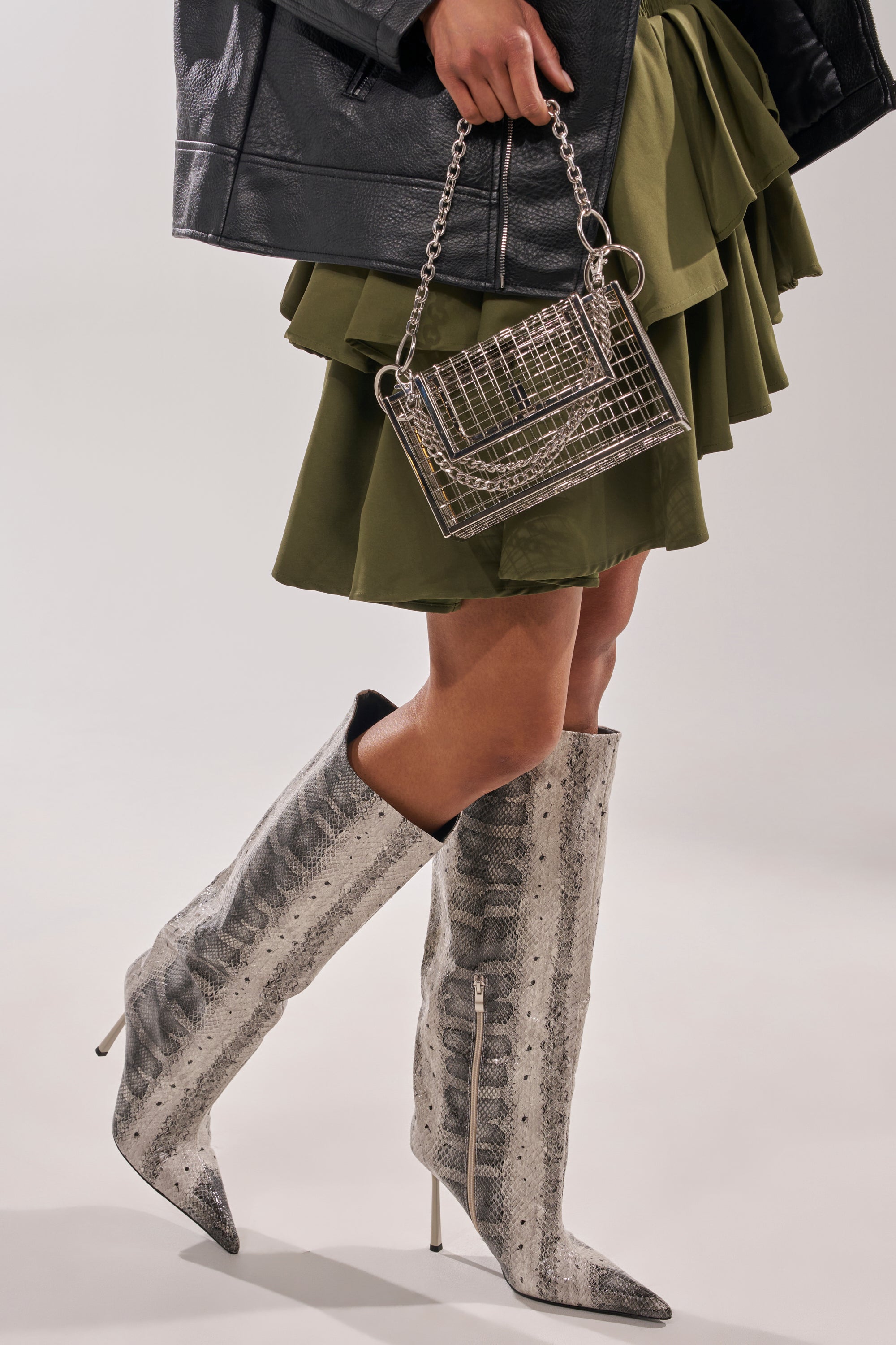 Wearing a dark leather jacket, olive green ruffled skirt, and AZALEA WANG MADEYE SNAKE STILETTO BOOTS, a person holds a metallic grid-patterned chain-strap handbag against a plain light background.