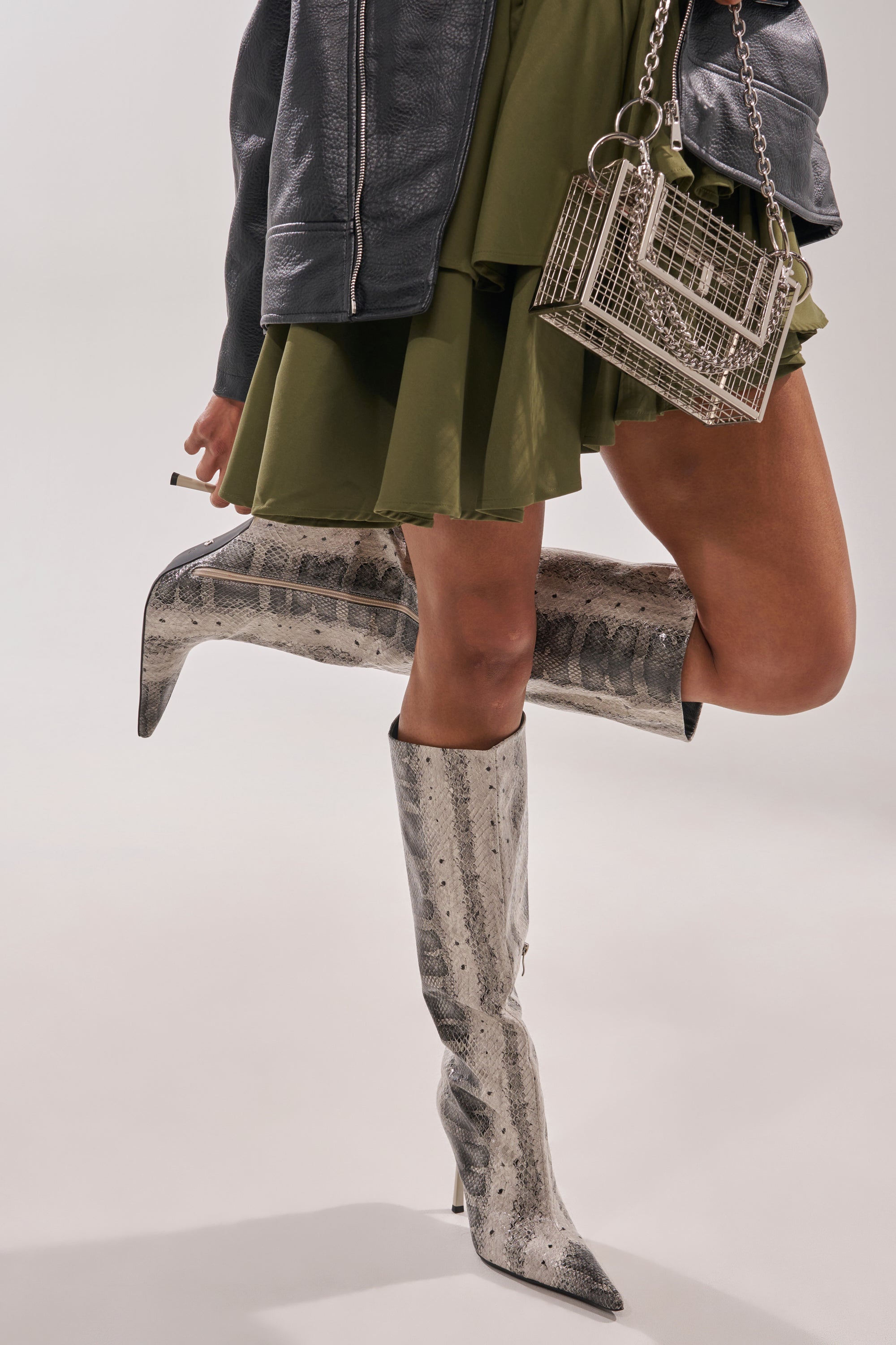 A person in a green skirt, textured gray jacket, and metallic grid handbag stands against a light background, styled with AZALEA WANG MADEYE SNAKE STILETTO BOOTS.