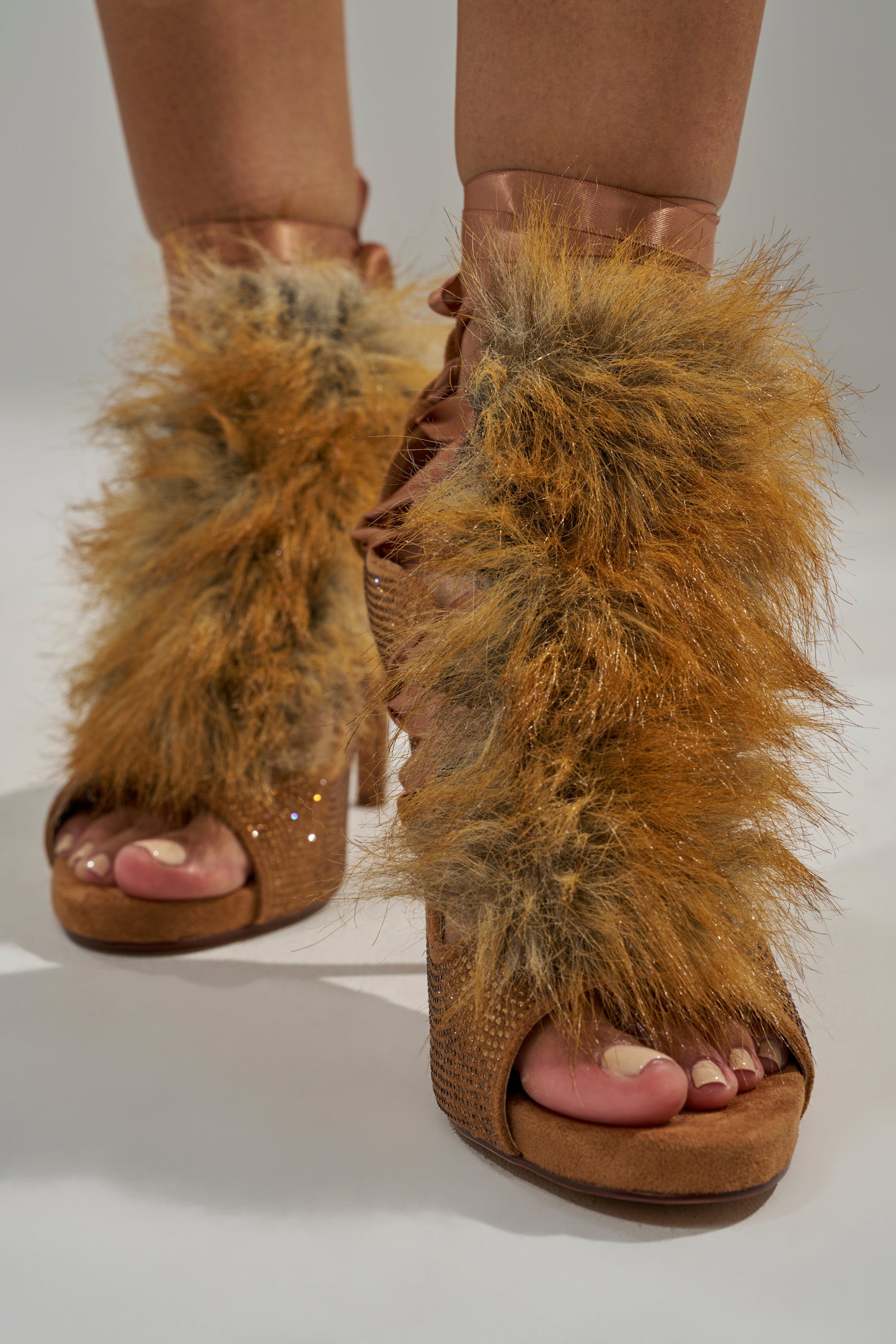 A close-up of a person wearing the AZALEA WANG MADHAV Bronze Rhinestone and Fox Fur Sandal, featuring thick brown and tan faux fur, an open toe, laced front, textured heels, and sparkling rhinestone details.