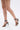 A person wearing AKIRA MADRIDI Black PVC and Metal Sandals with stiletto heels and silver oval-accented ankle straps, walking against a plain white background.
