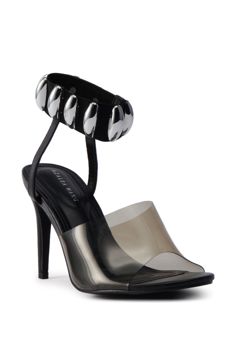 The AKIRA MADRIDI BLACK PVC AND METAL SANDAL features a stiletto heel, a clear PVC toe strap, and a metal-accented ankle strap adorned with large silver oval embellishments. 4