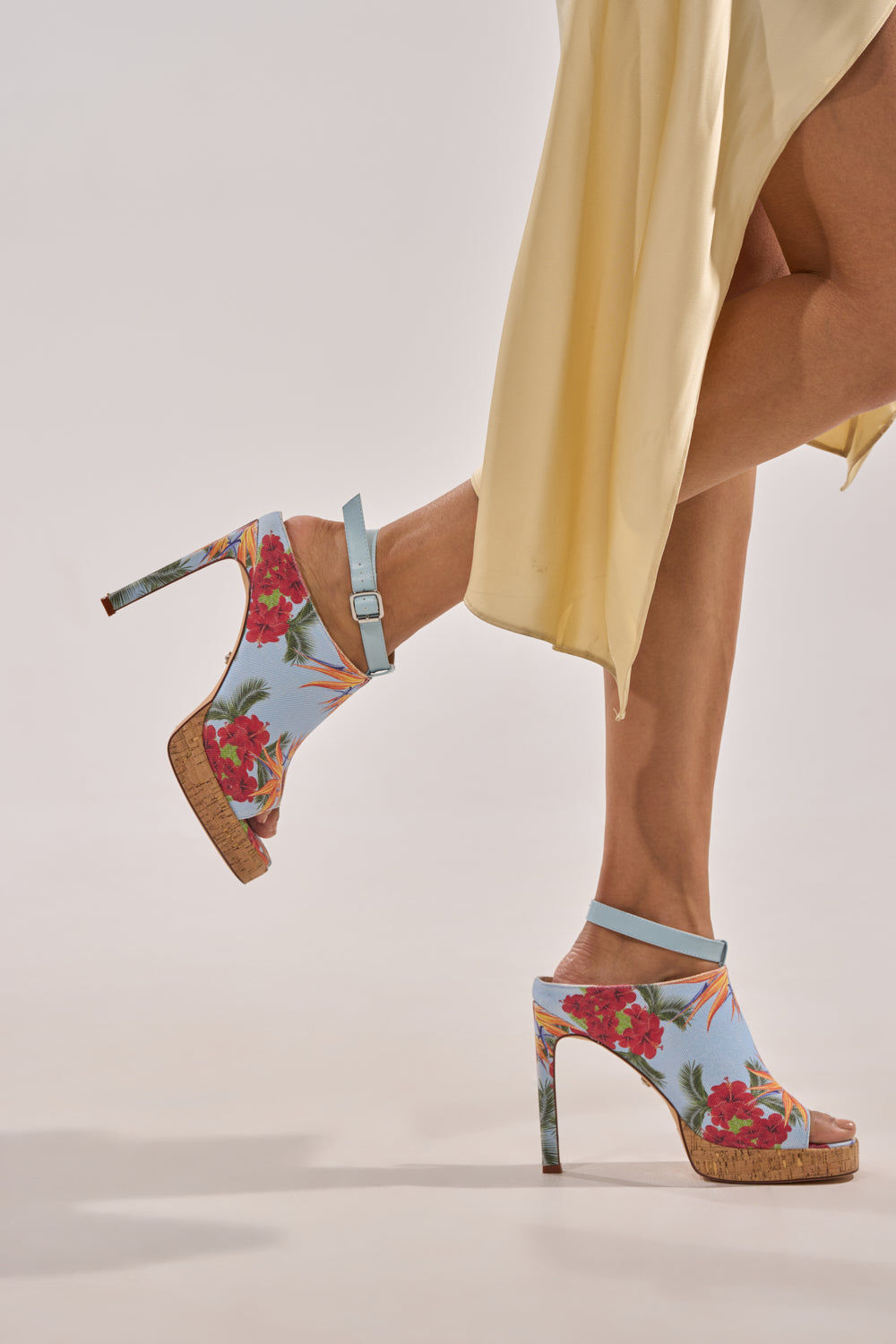 A person in a flowing yellow dress walks with one leg raised, wearing AZALEA WANG MAELIE MULTI FLORAL STILETTO SANDALS featuring a blue tropical print, ankle straps, and cork soles. 1