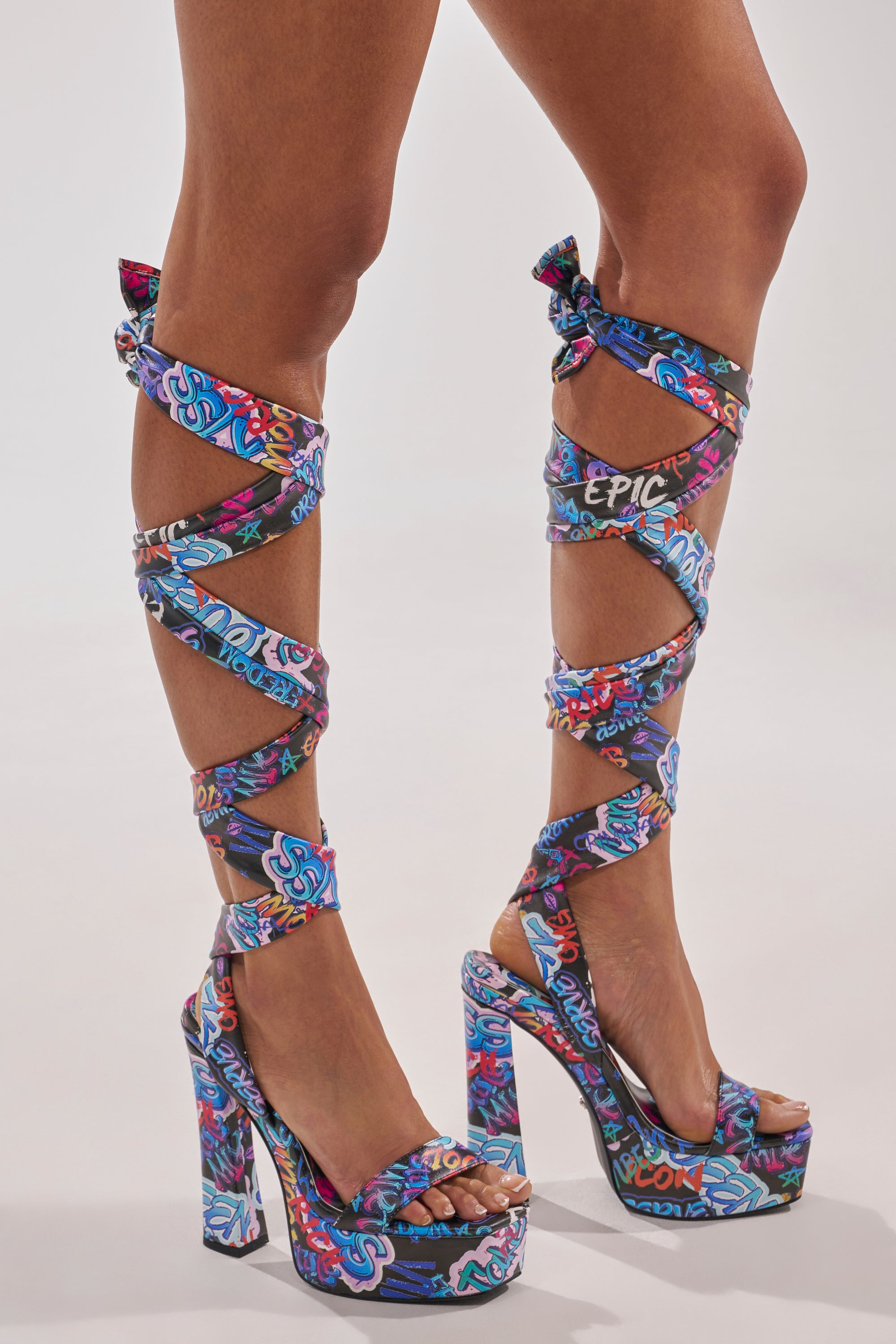 A person wears the AKIRA GOLD MAGRITTE GRAFFITI STRAPPY CHUNKY SANDAL—platform sandals with colorful graffiti print and matching straps winding up their calves, tied in bows below the knees, making a bold streetwear statement against a plain background. 0