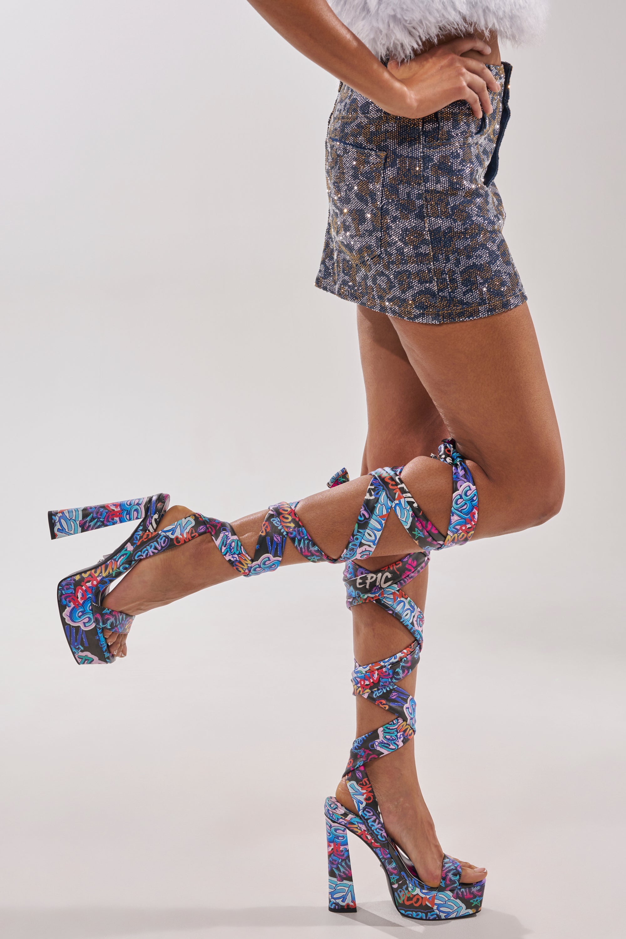 Wearing the AKIRA GOLD MAGRITTE GRAFFITI STRAPPY CHUNKY SANDAL and sequined shorts, a person stands with one leg raised against a plain background, showcasing the sandals’ colorful wraparound straps. 7