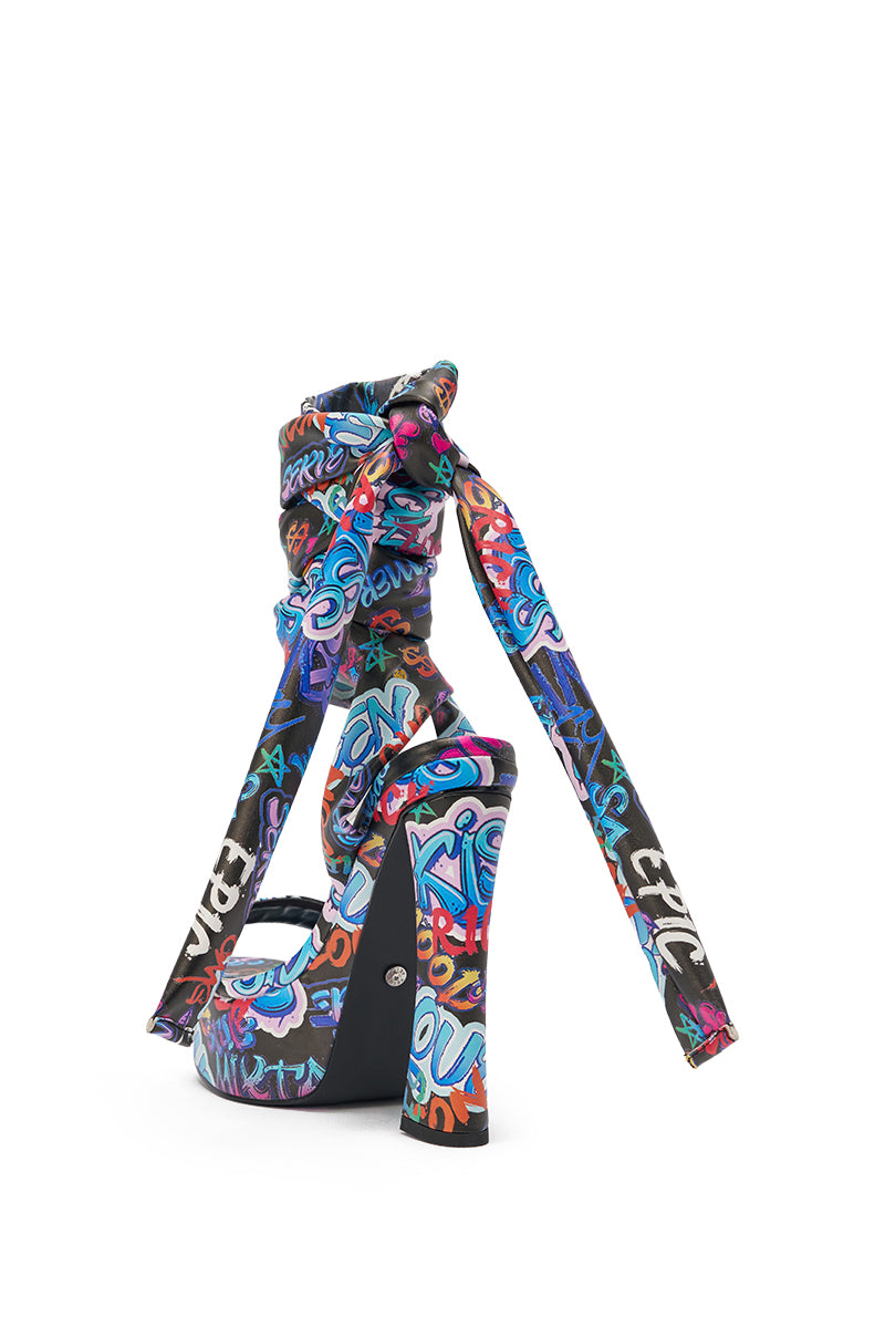The AKIRA GOLD MAGRITTE GRAFFITI STRAPPY CHUNKY SANDAL features multicolored graffiti-style text, a thick ankle-wrap bow strap, and a chunky heel—making it a bold statement piece against a crisp white background. 5