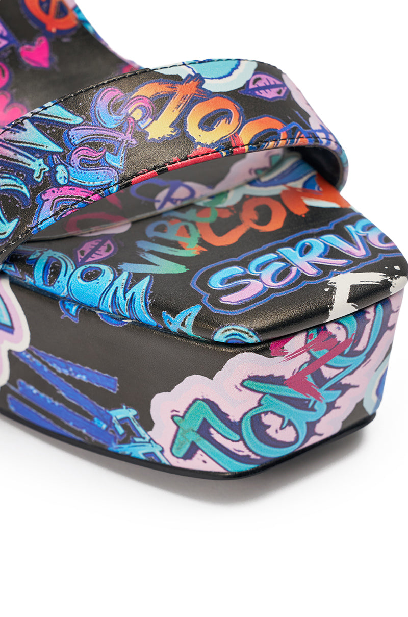 Close-up of the AKIRA GOLD MAGRITTE GRAFFITI STRAPPY CHUNKY SANDAL, featuring colorful street-style text in blue, purple, orange, and pink on black. These statement shoes have a thick sole and a toe strap. 6