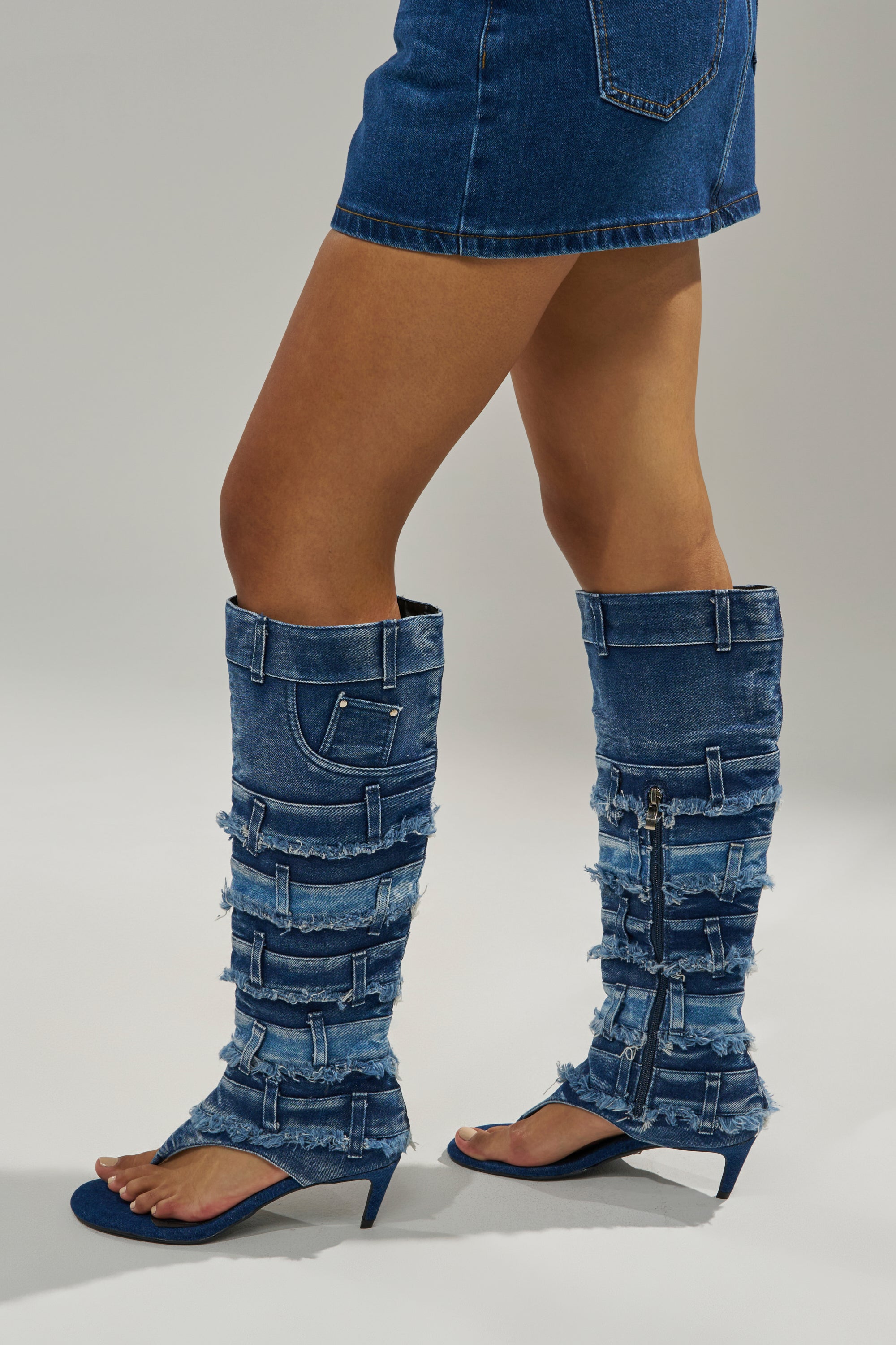 A person wearing a short denim skirt and AKIRA GOLD MAHONEY DENIM PANT DETAIL GLADIATOR STILETTO heels stands against a plain background. 0