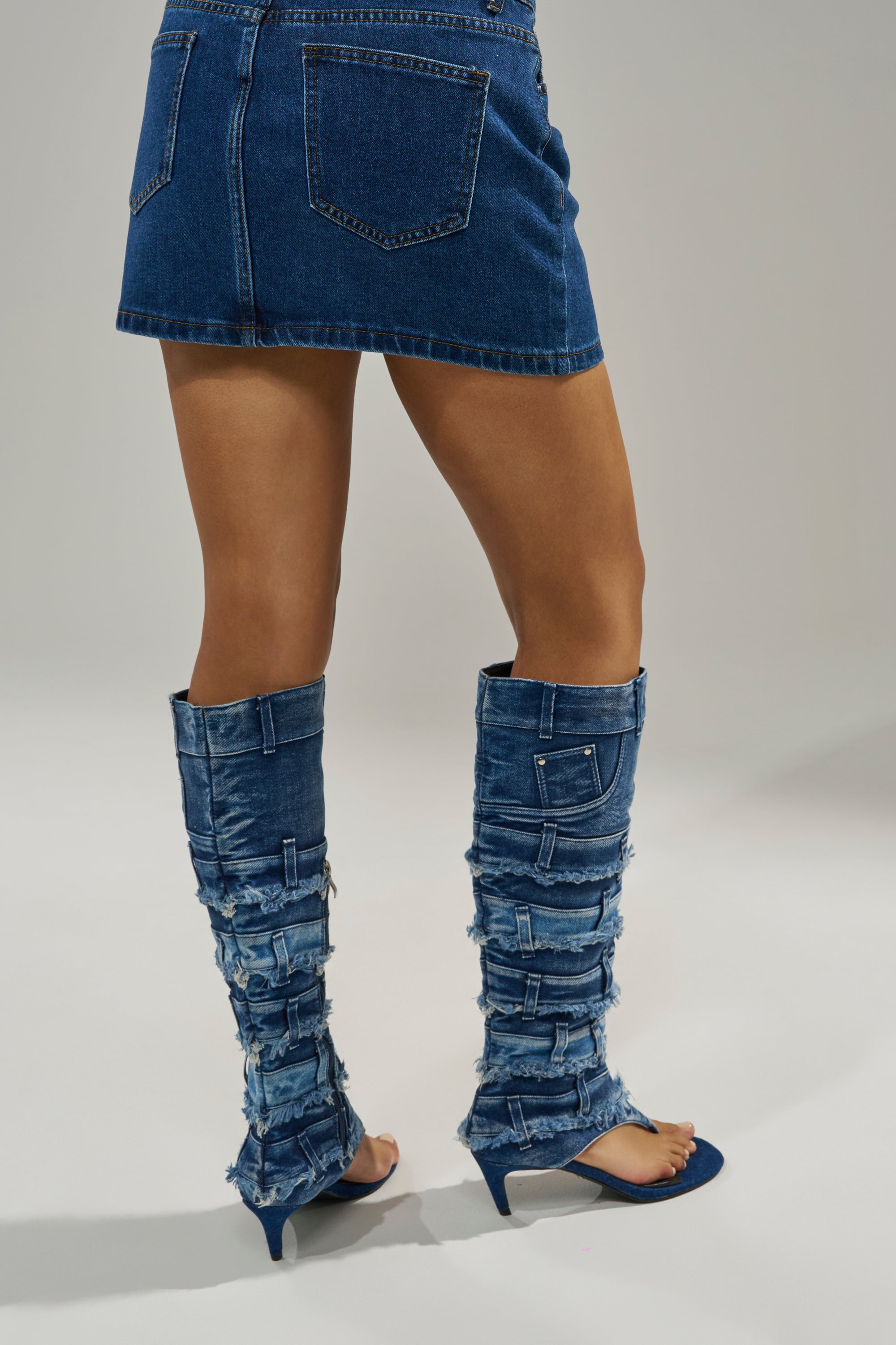 Wearing a short blue denim skirt and the AKIRA GOLD MAHONEY DENIM PANT DETAIL GLADIATOR STILETTO with open toes and distressed denim heels, a person poses against a plain light gray background. 2